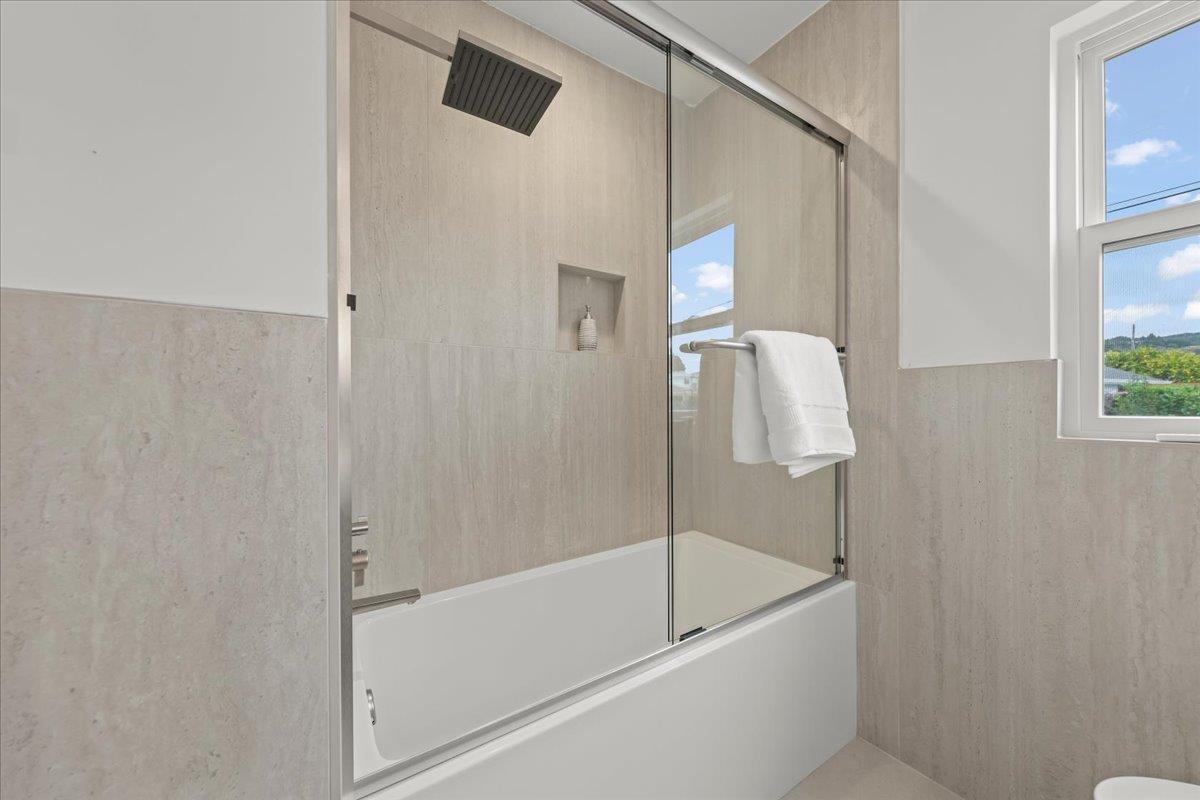 Bathroom with a glass-enclosed bathtub, beige tile, and a window overlooking greenery.