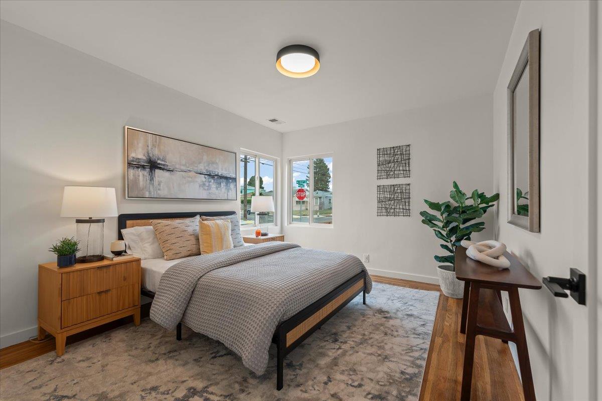 Staged bedroom with a large gray-covered bed, wood nightstand, and street-facing windows.