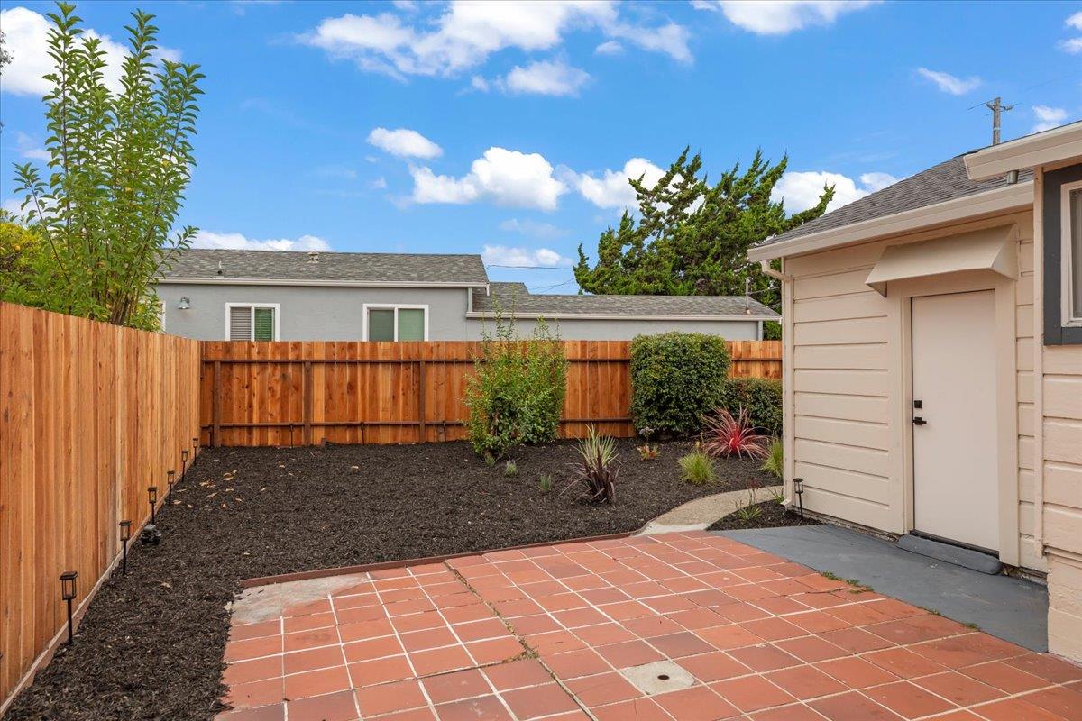 Fenced backyard with a terracotta-tiled patio, planted beds, and a small beige outbuilding.
