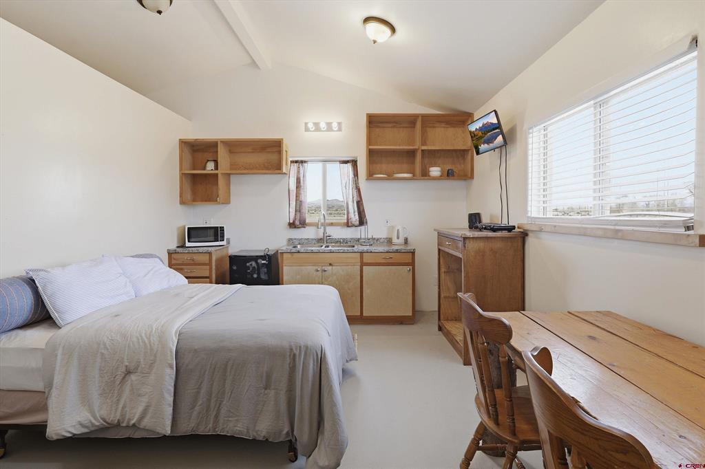 Bedroom with a kitchenette, wooden dining table, and large window with white blinds.