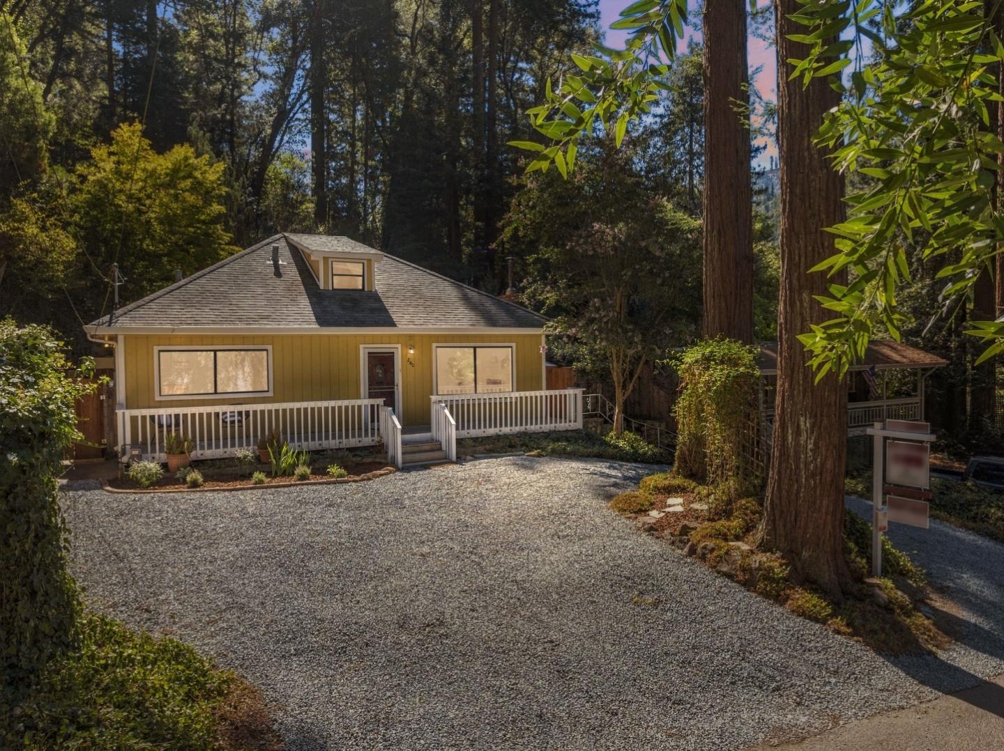 Yellow house with a white-railed porch and gravel driveway among tall redwood trees.