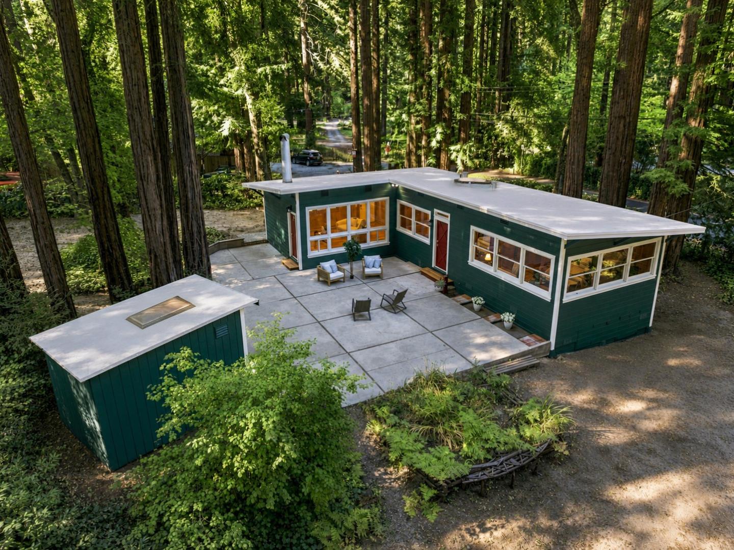 Modern green home with a broad patio and matching outbuilding nestled among tall trees.