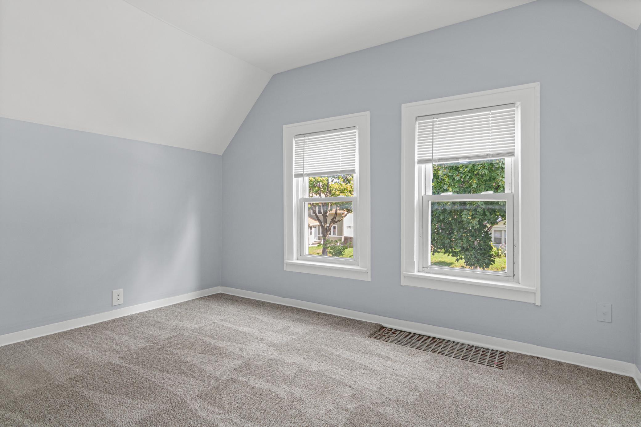 Empty carpeted room with a sloped ceiling, two windows, and a long floor vent.