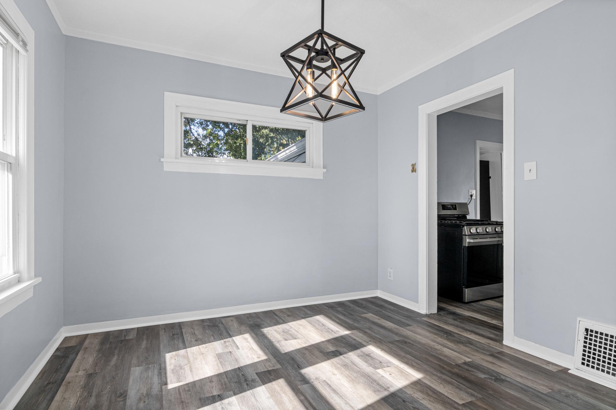 Empty gray room with a geometric pendant light, sunlit plank flooring, and a view into the kitchen.