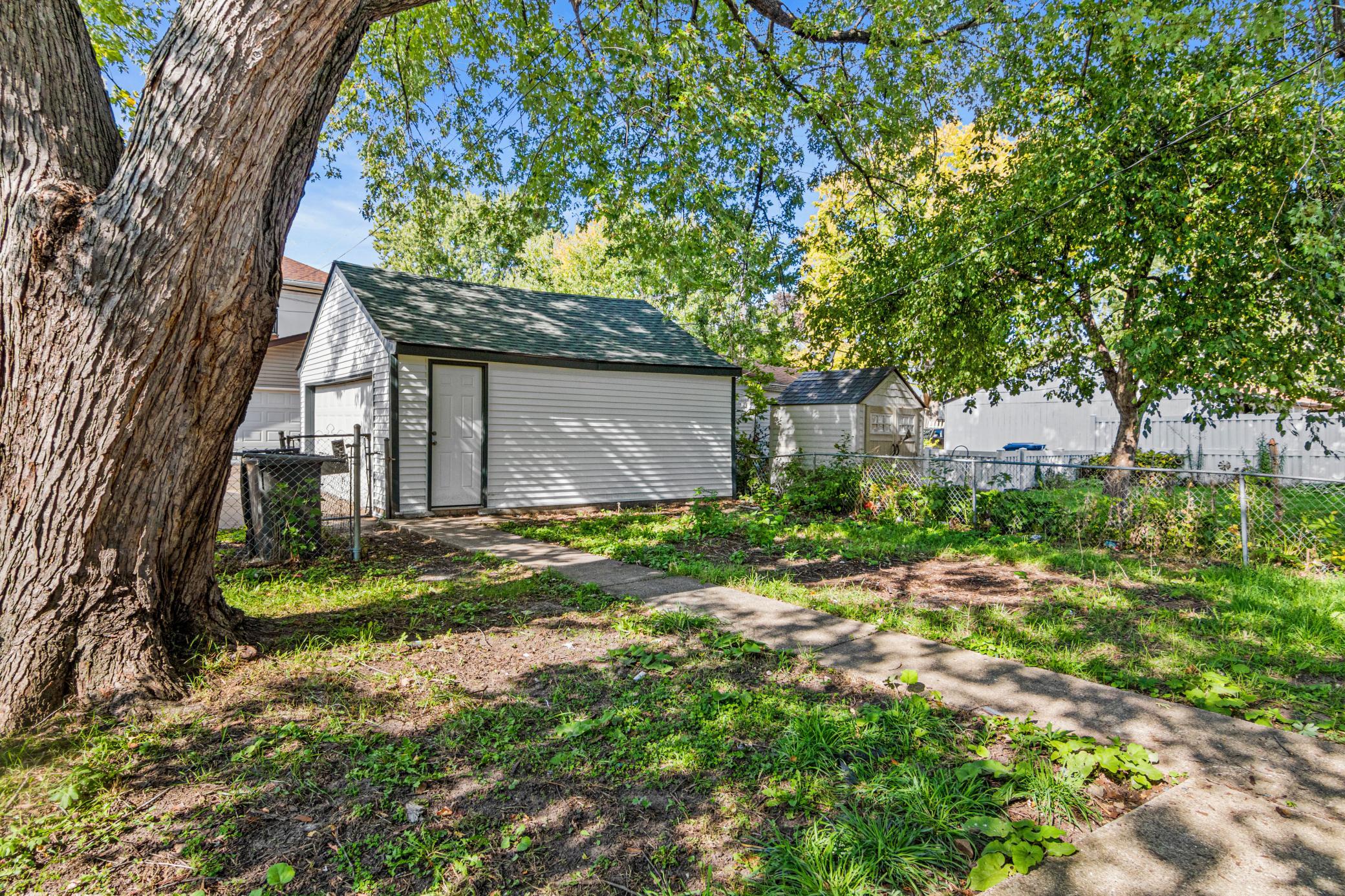Detached gray garage with a side door, shaded by a large tree beside a concrete path.