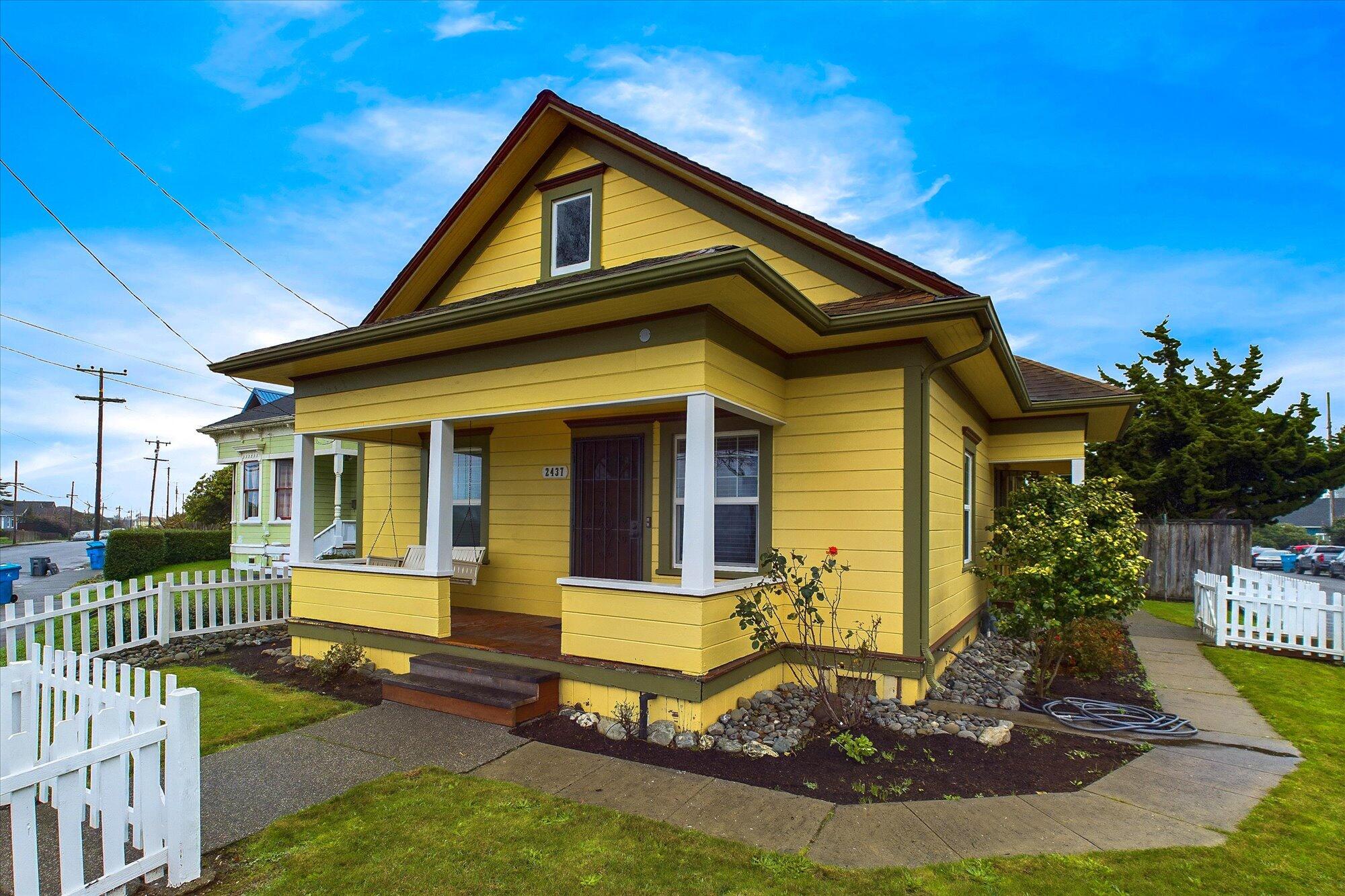 Yellow house with a covered porch, white picket fence, and landscaped front yard.