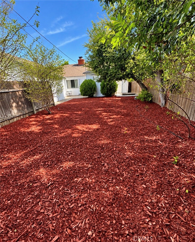 Fenced backyard covered in red mulch, with leafy trees and a white house beyond.
