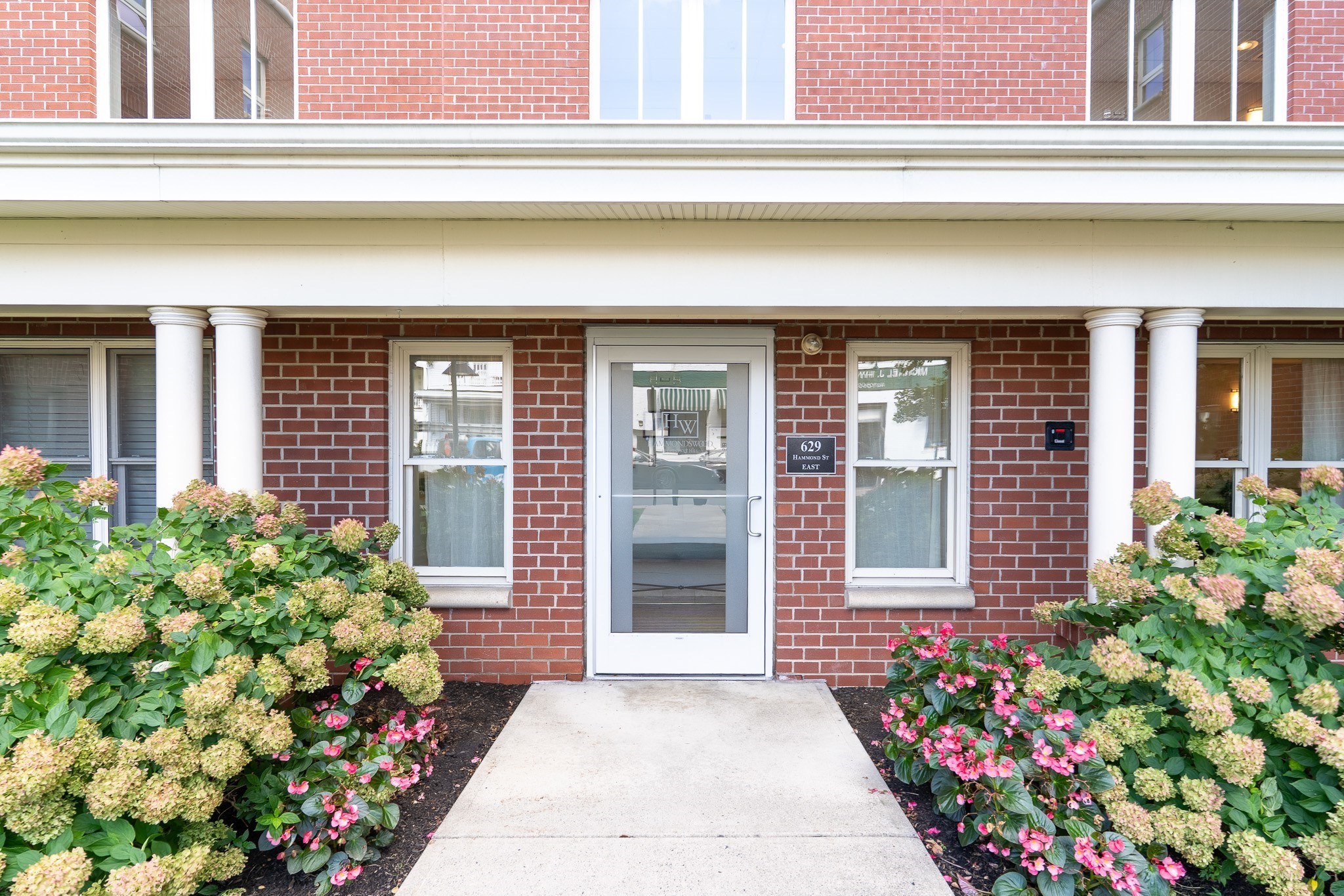 Brick building entrance with white columns, a glass door, and flowering shrubs.