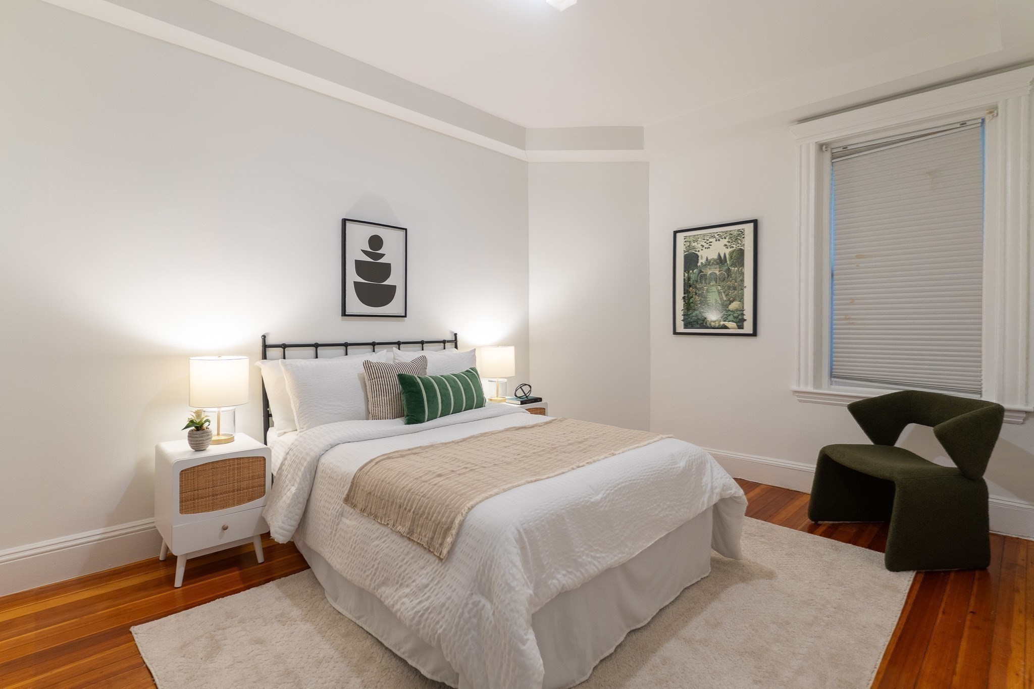 Staged bedroom with a white bed, green accent pillows, hardwood floors, and a dark green chair.
