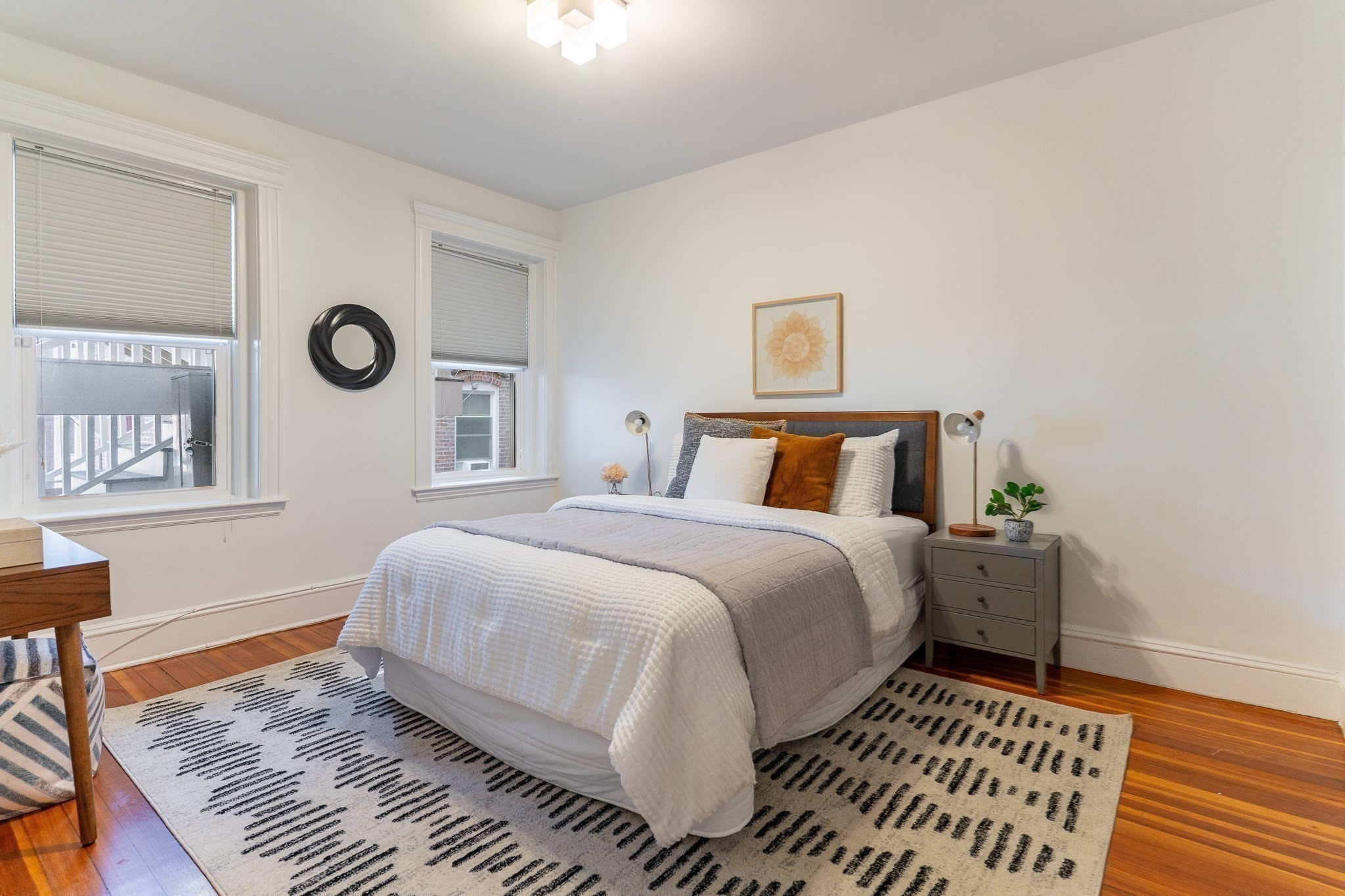Staged bedroom with a gray upholstered bed, patterned rug, hardwood floors, and two windows.
