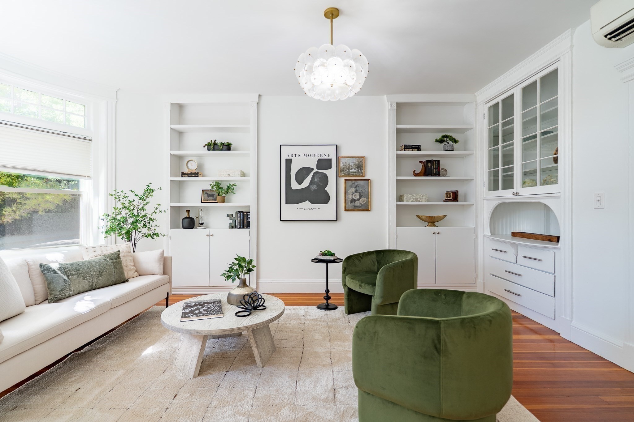Bright living room with white built-ins, a cream sofa, green armchairs, and a sculptural pendant light.