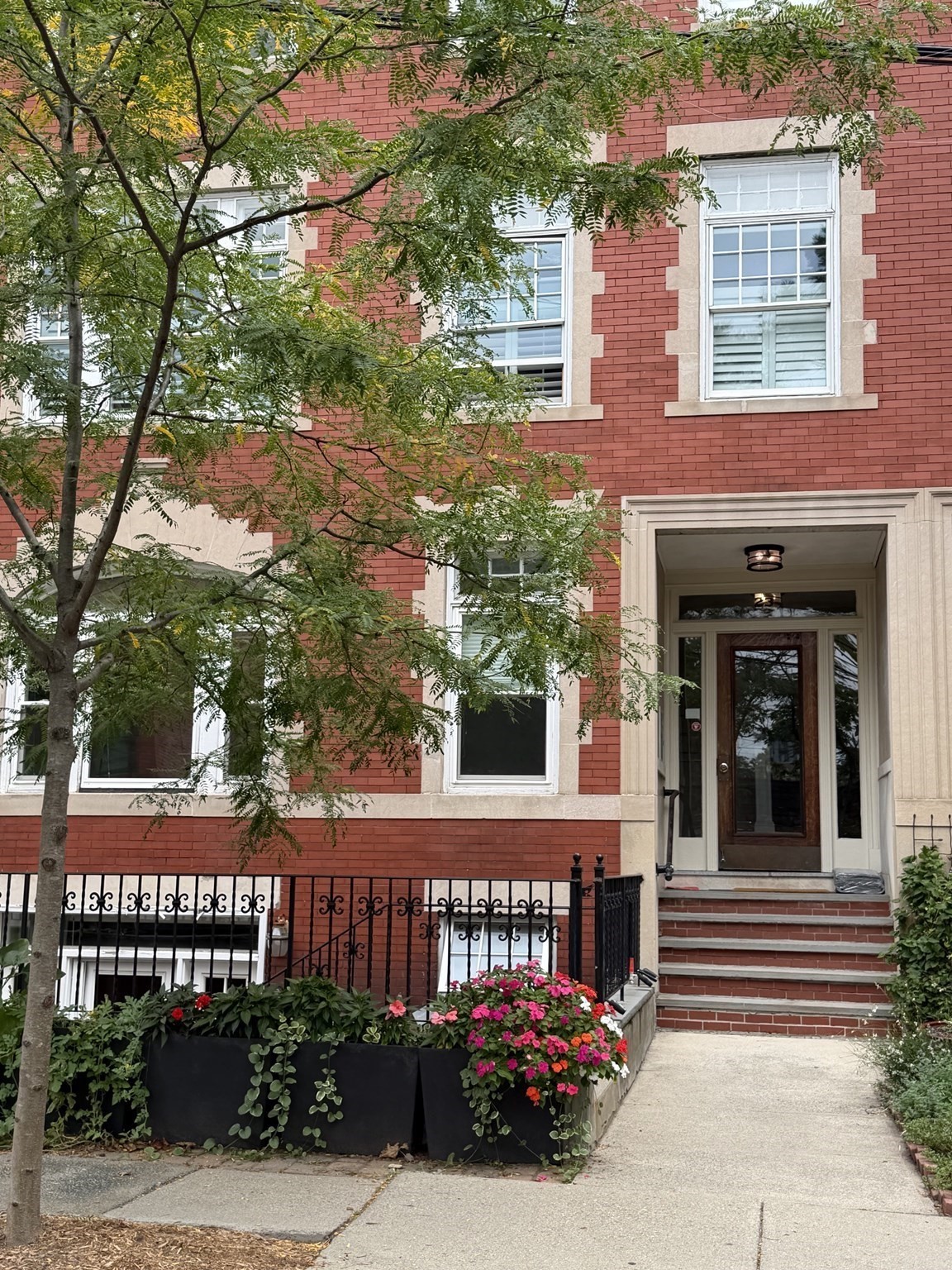 Red-brick residence entrance with stone trim, front steps, and flower planters beneath leafy trees.