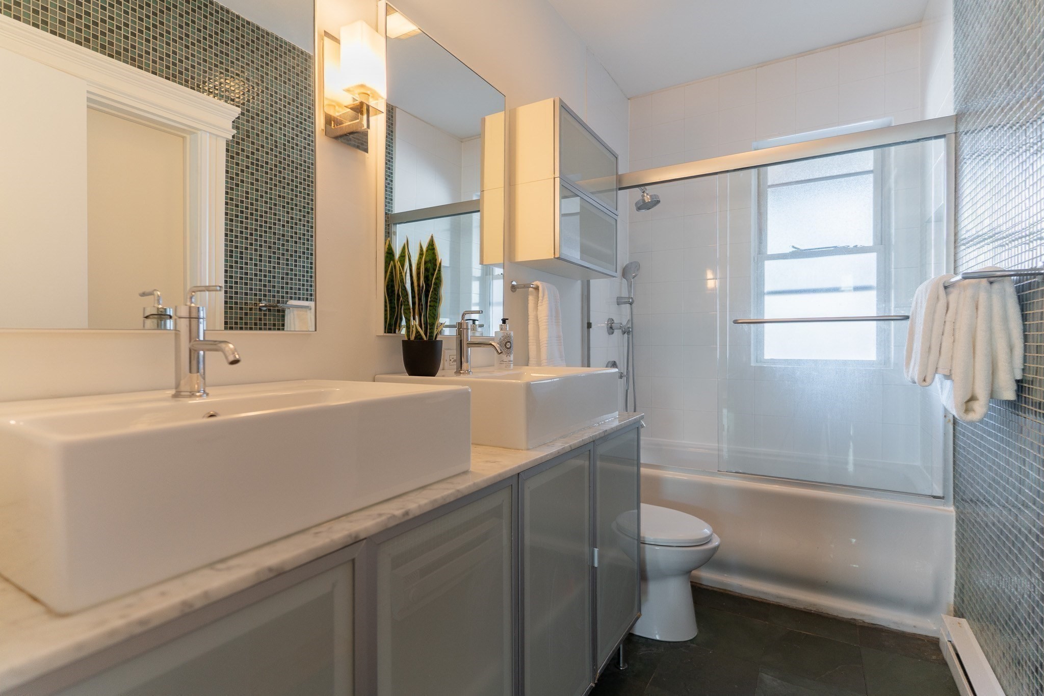 Modern bathroom with double vessel sinks, dark mosaic tile accents, and a glass-enclosed tub and shower.