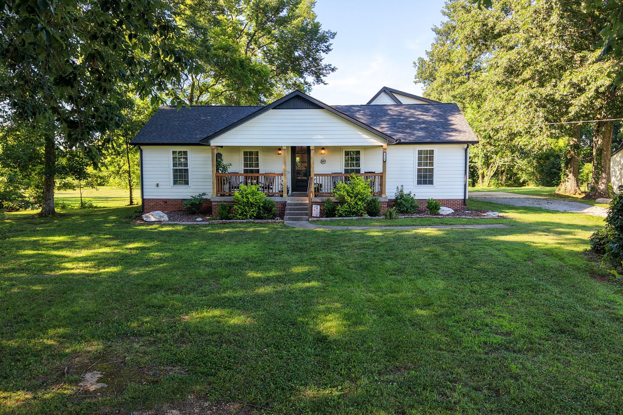 White single-story house with a covered porch, brick foundation, and mature trees around the lawn.