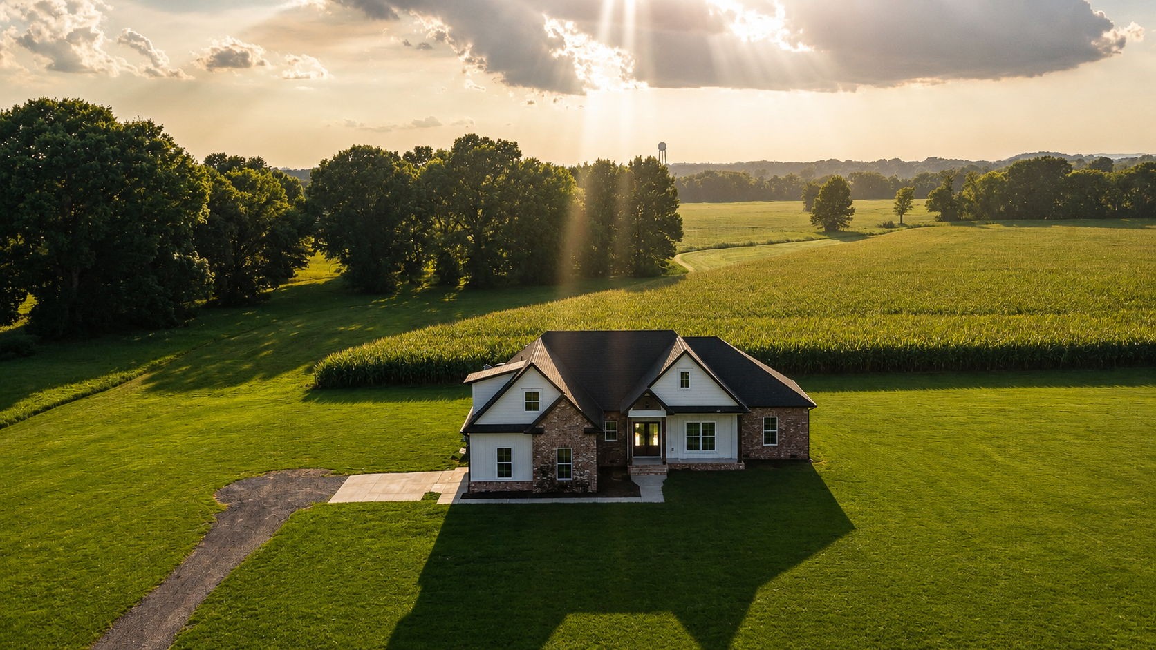 Aerial view of a brick-and-white house amid broad green lawns, fields, and trees at golden hour.