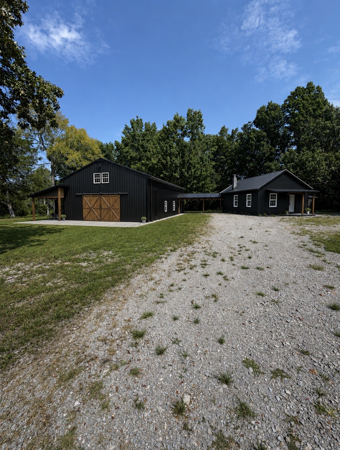 Black farmhouse beside a matching barn-style outbuilding, with a wide gravel driveway and wooded surroundings.