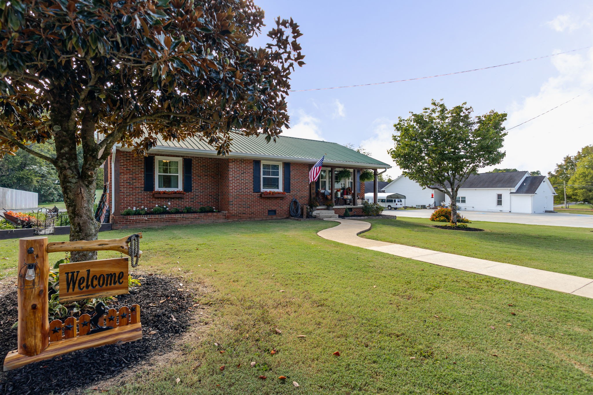 Red brick home with green metal roof, covered porch, curved walkway, and wooden welcome sign.