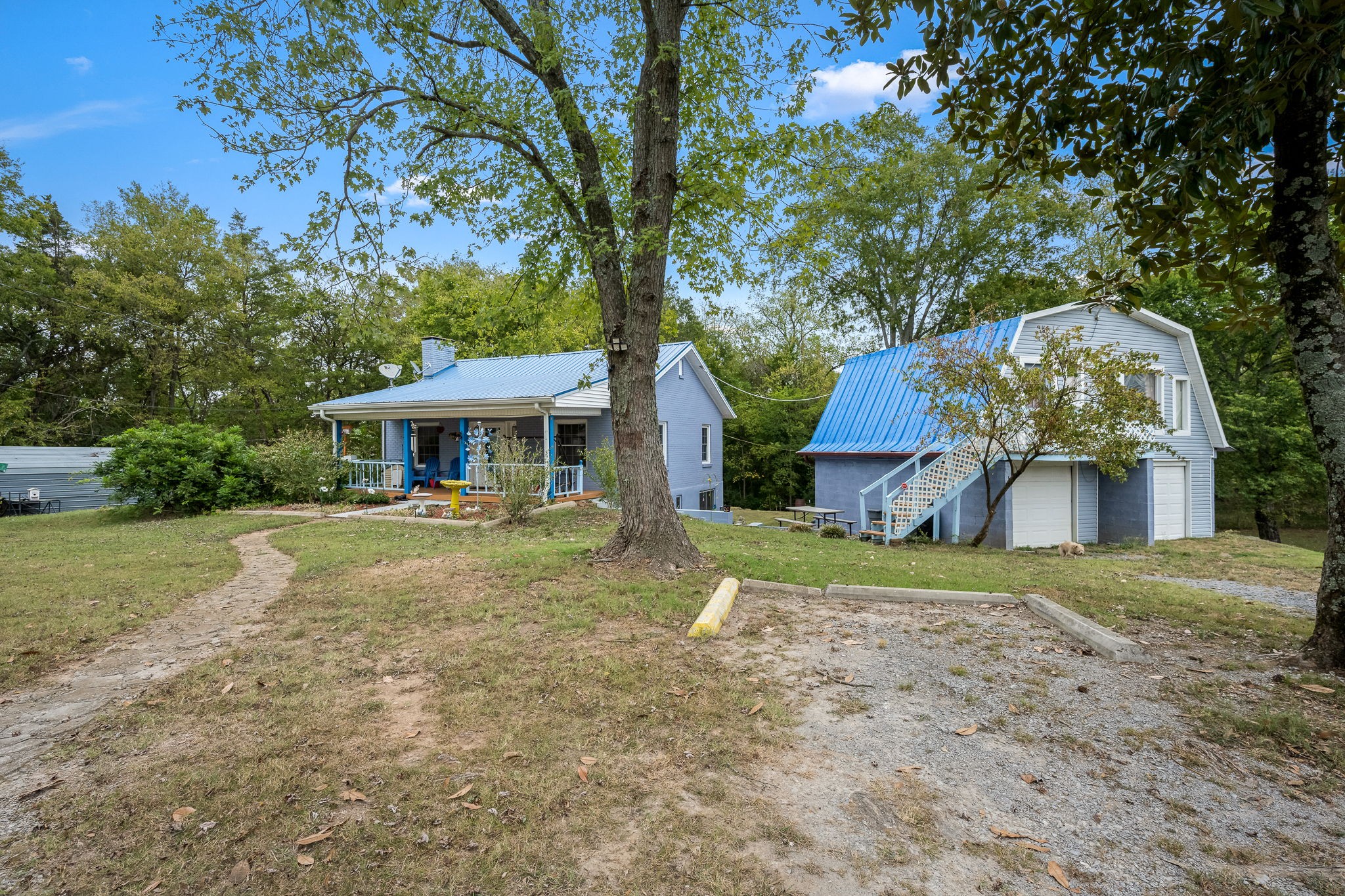 Blue-roofed house and larger outbuilding among mature trees, with a gravel parking area and stone path.
