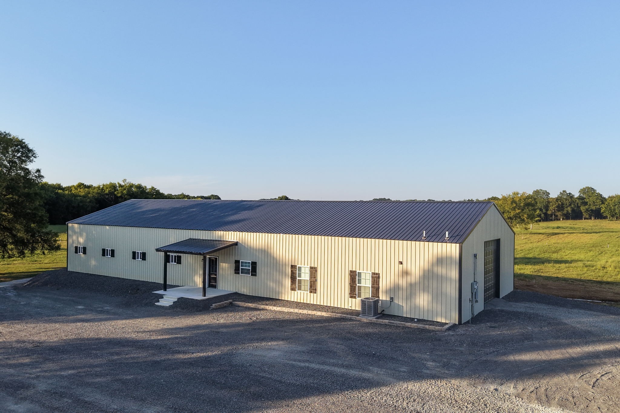 Large metal building with a covered entrance and garage door beside open grassy fields.