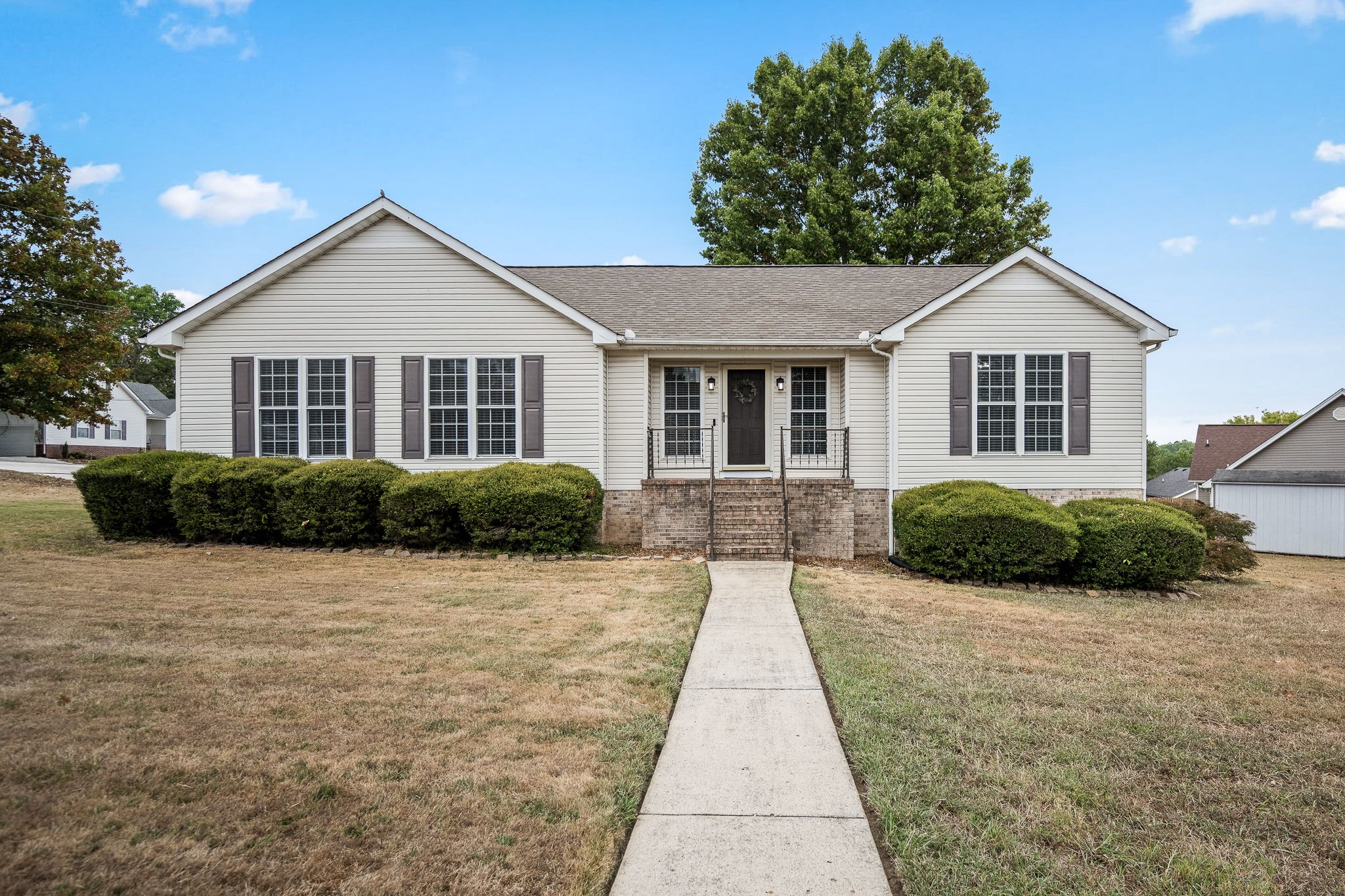 Single-story beige house with front gables, brick steps, trimmed shrubs, and a concrete walkway.