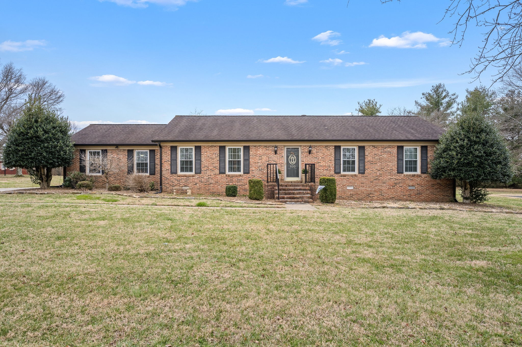 Single-story brick ranch house with a central entrance, flanking shrubs, and a broad lawn.