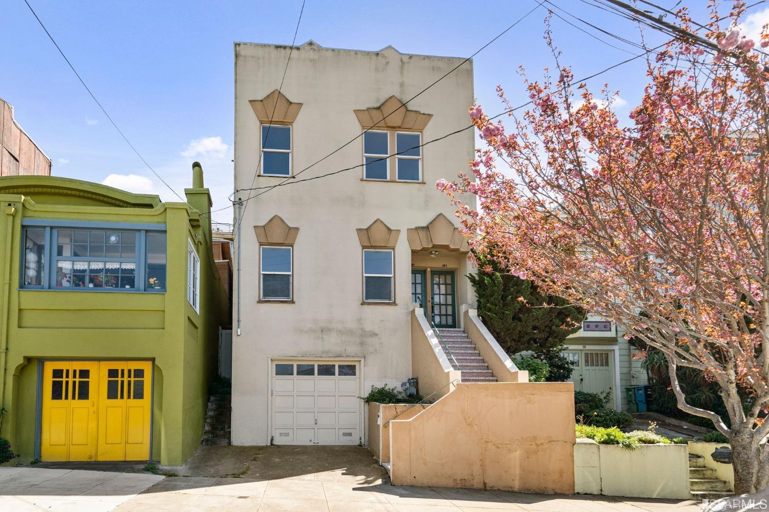 Cream three-story home with geometric window details, front steps, garage, and a pink-blossoming tree.