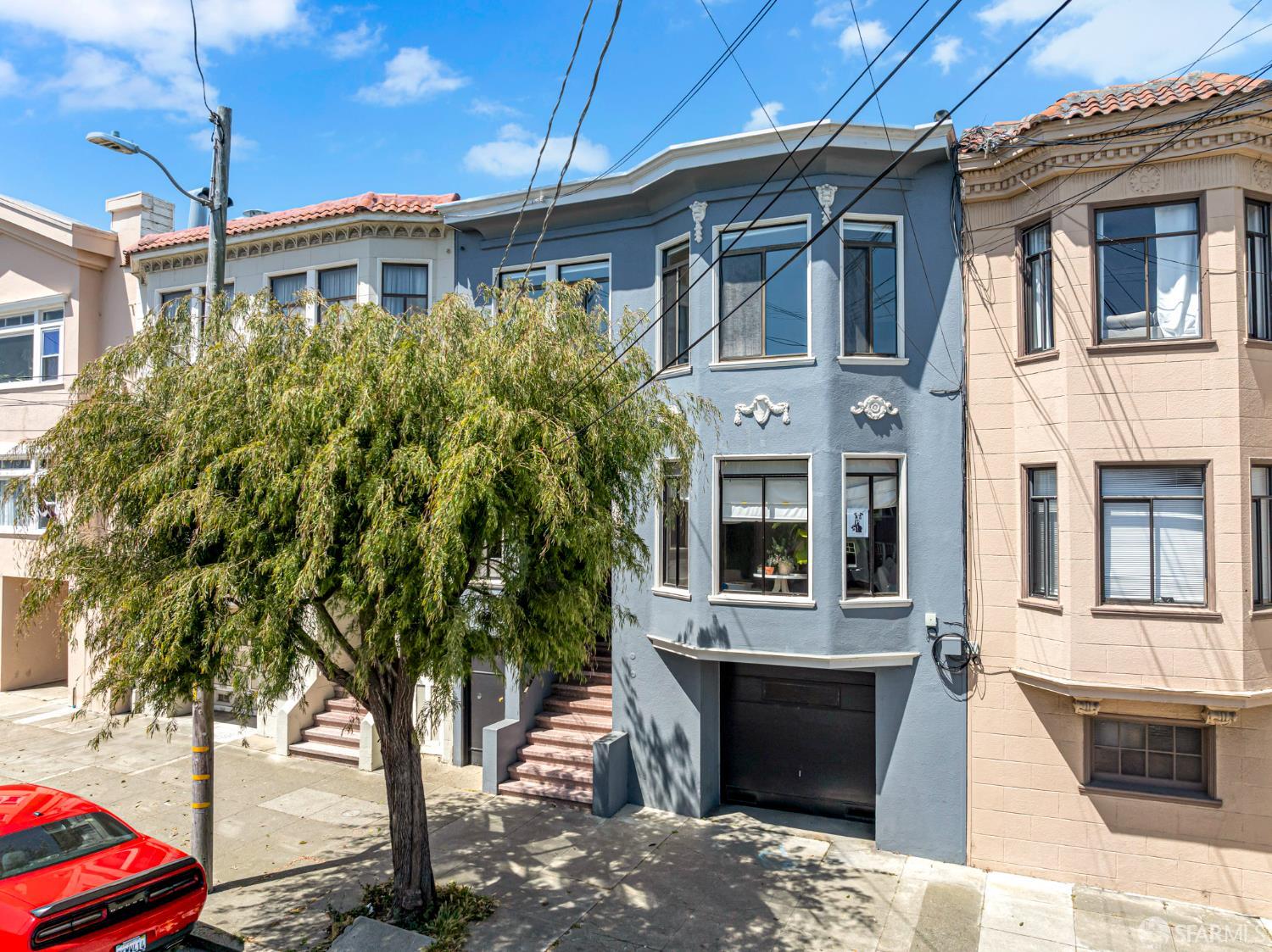 Blue multi-story home with white trim and a garage, partly shaded by a leafy street tree.