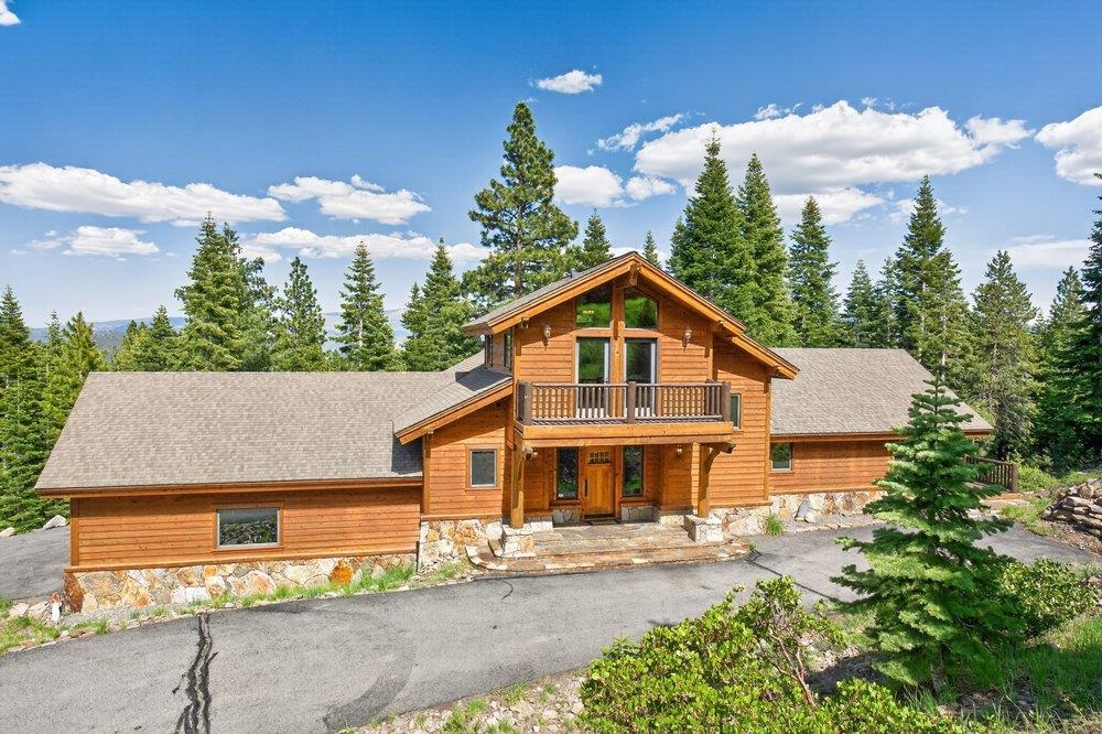 Wood-sided mountain home with a stone foundation and upper balcony, surrounded by pine trees.