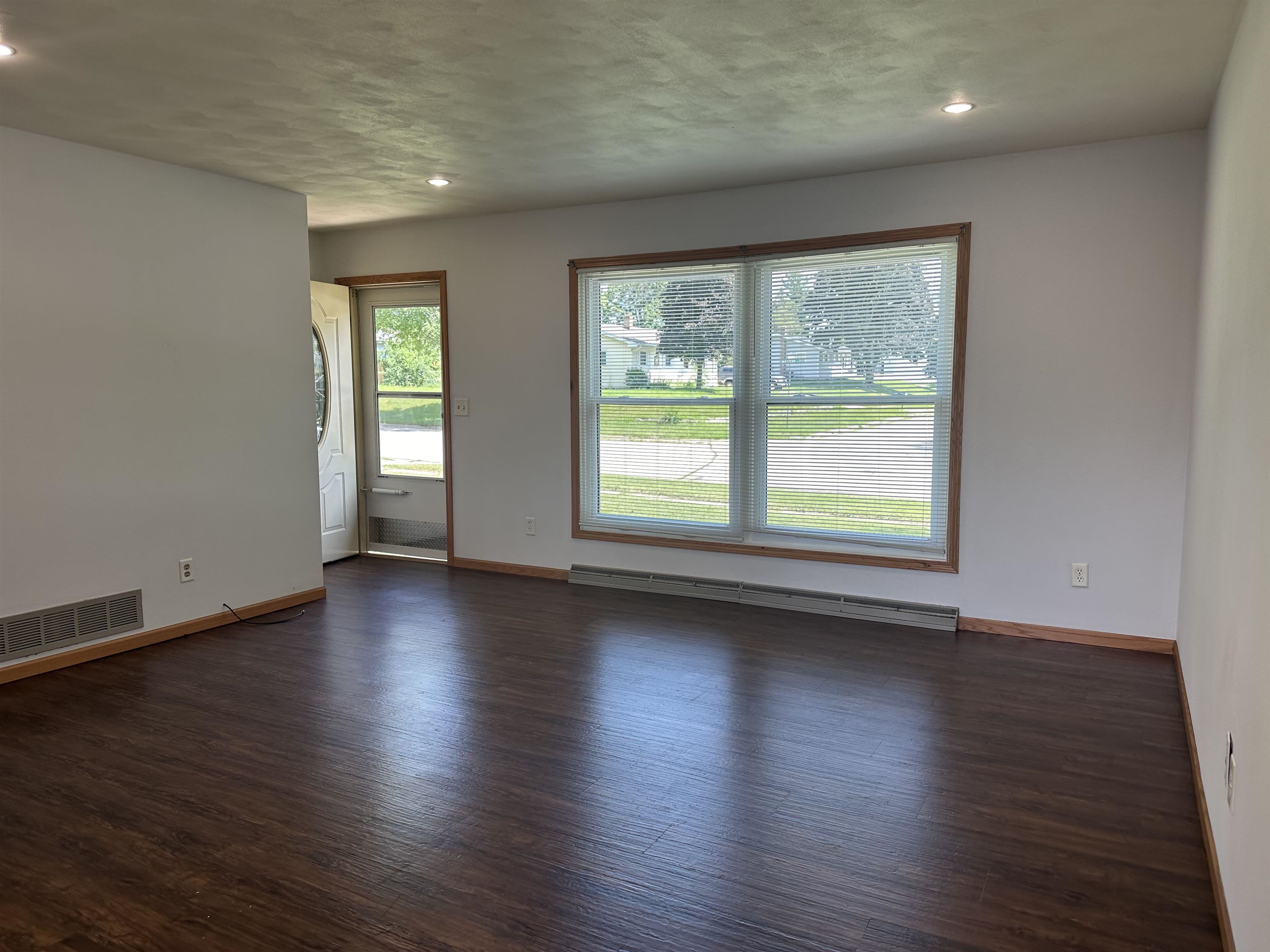 Vacant living room with dark plank flooring and a large front window with blinds.