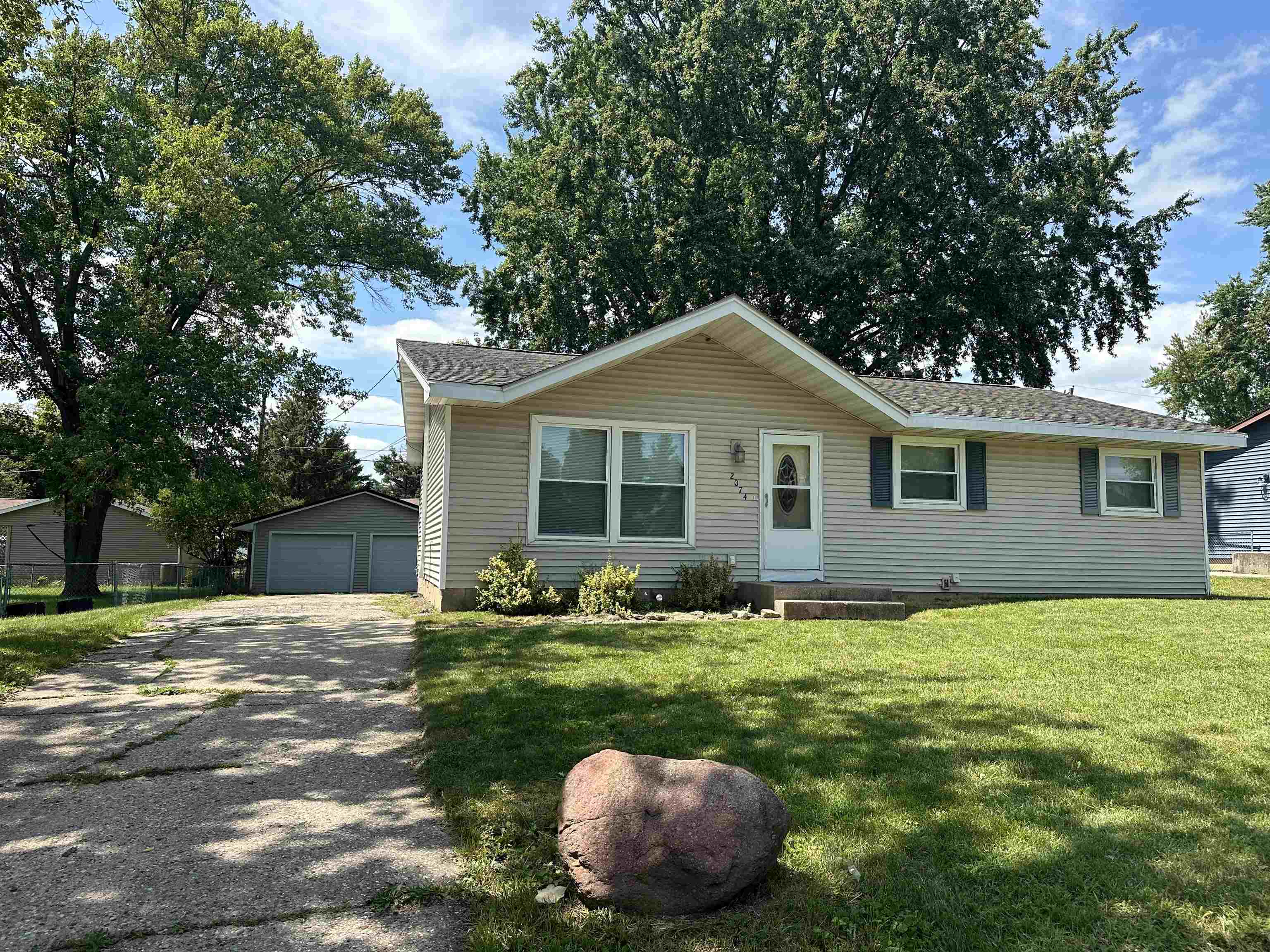 Single-story beige home with a detached garage, concrete driveway, and mature trees.