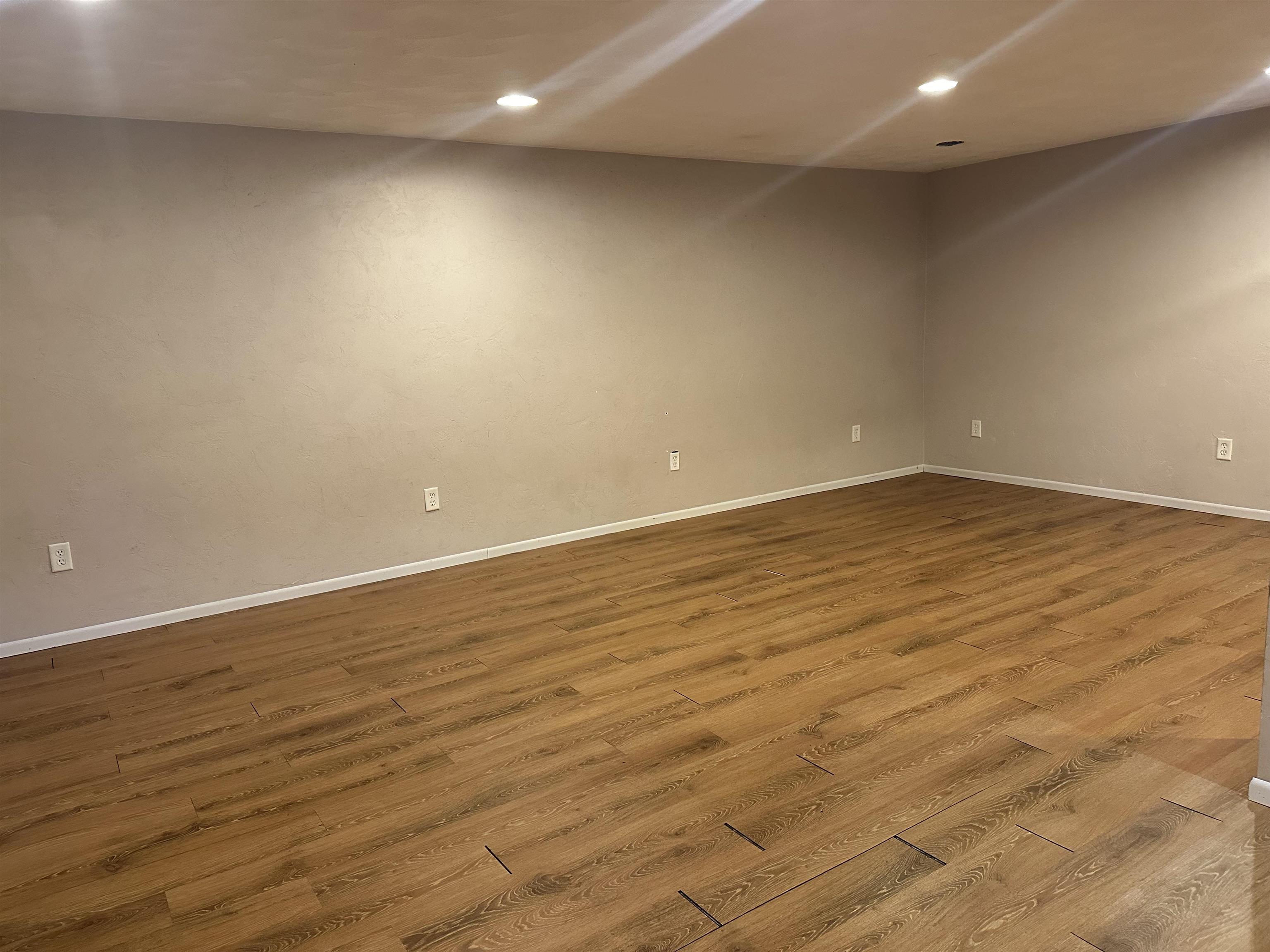 Empty room with warm wood-plank flooring, beige walls, and recessed ceiling lights.