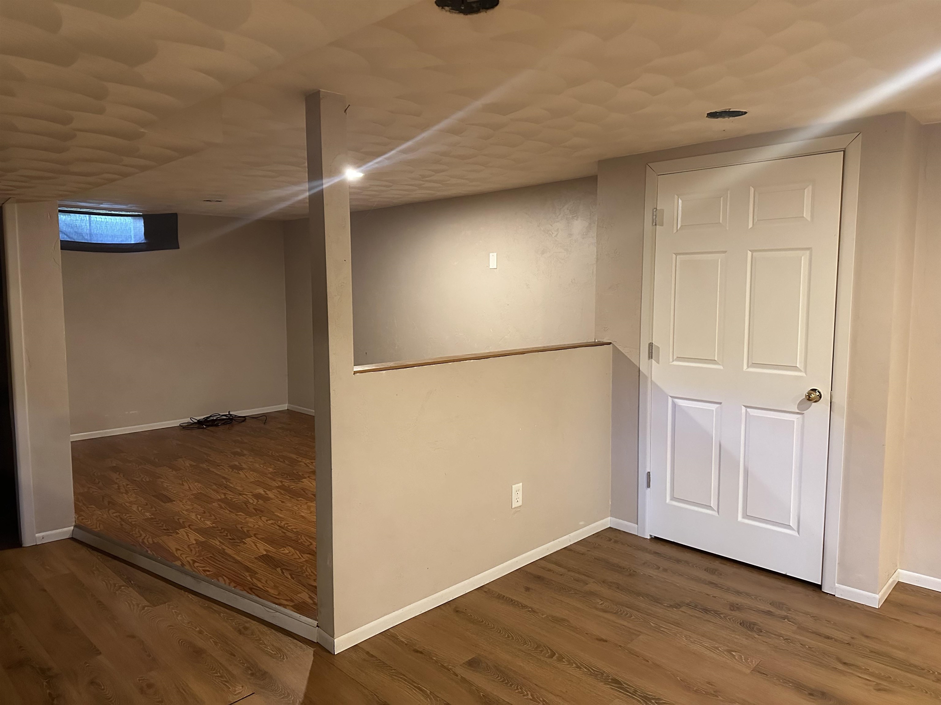 Empty basement room with wood-look flooring, a half wall, and a small high window.