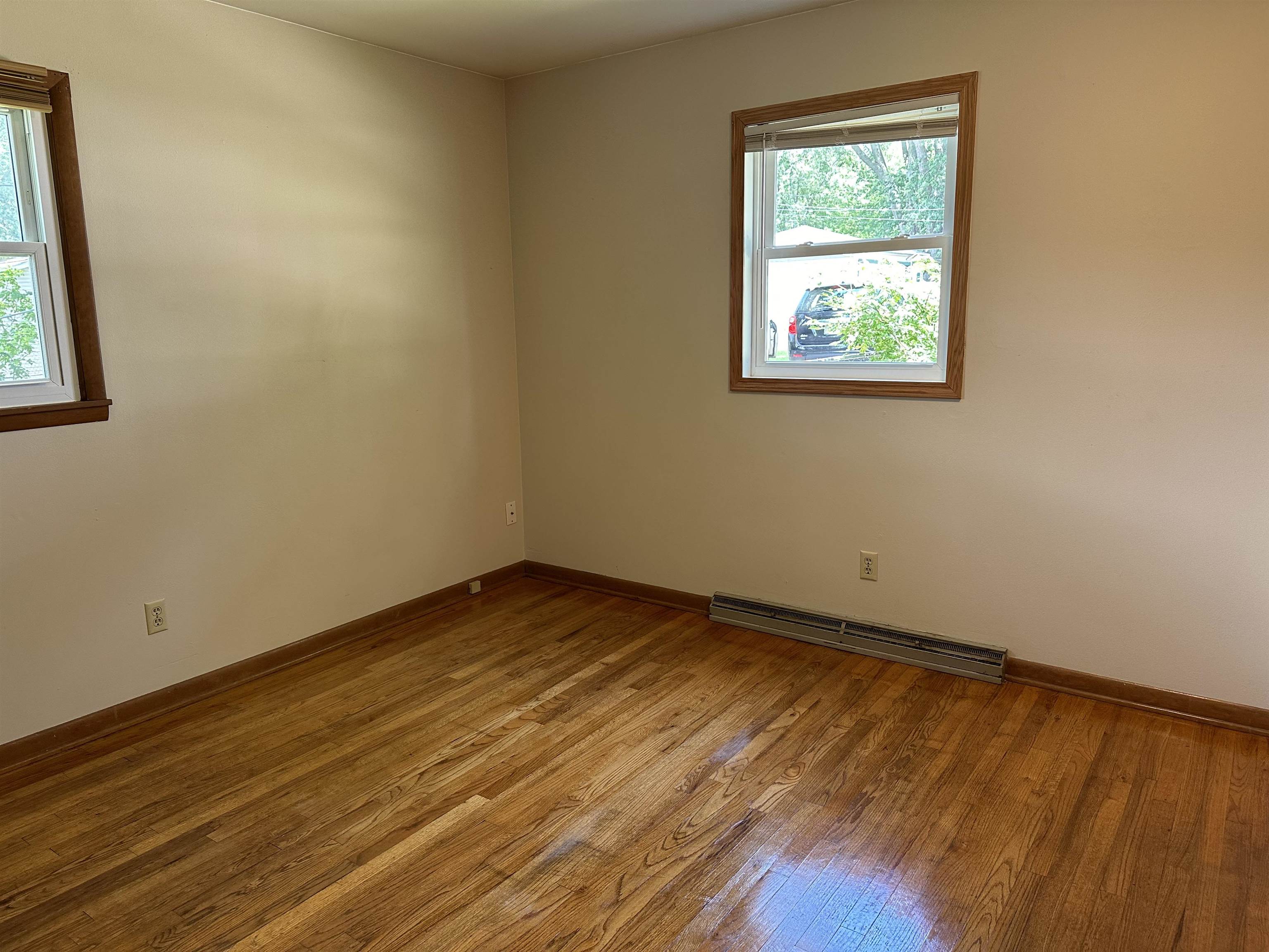 Empty room with polished hardwood floors, two windows, and a baseboard heater.