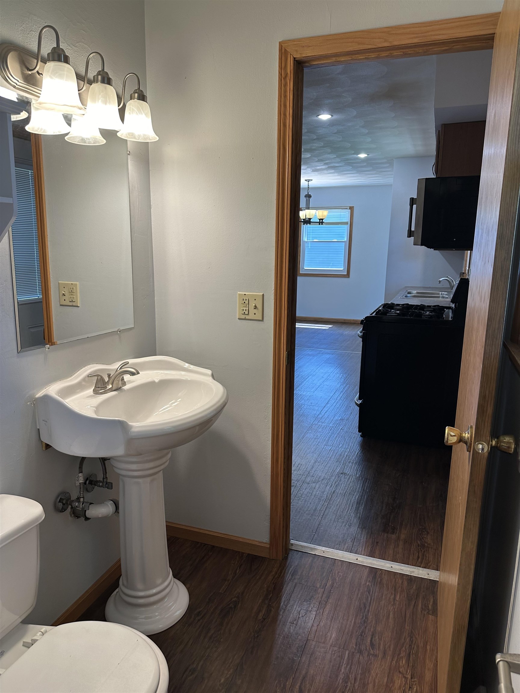 Bathroom with pedestal sink and mirror, looking through a doorway into the kitchen.