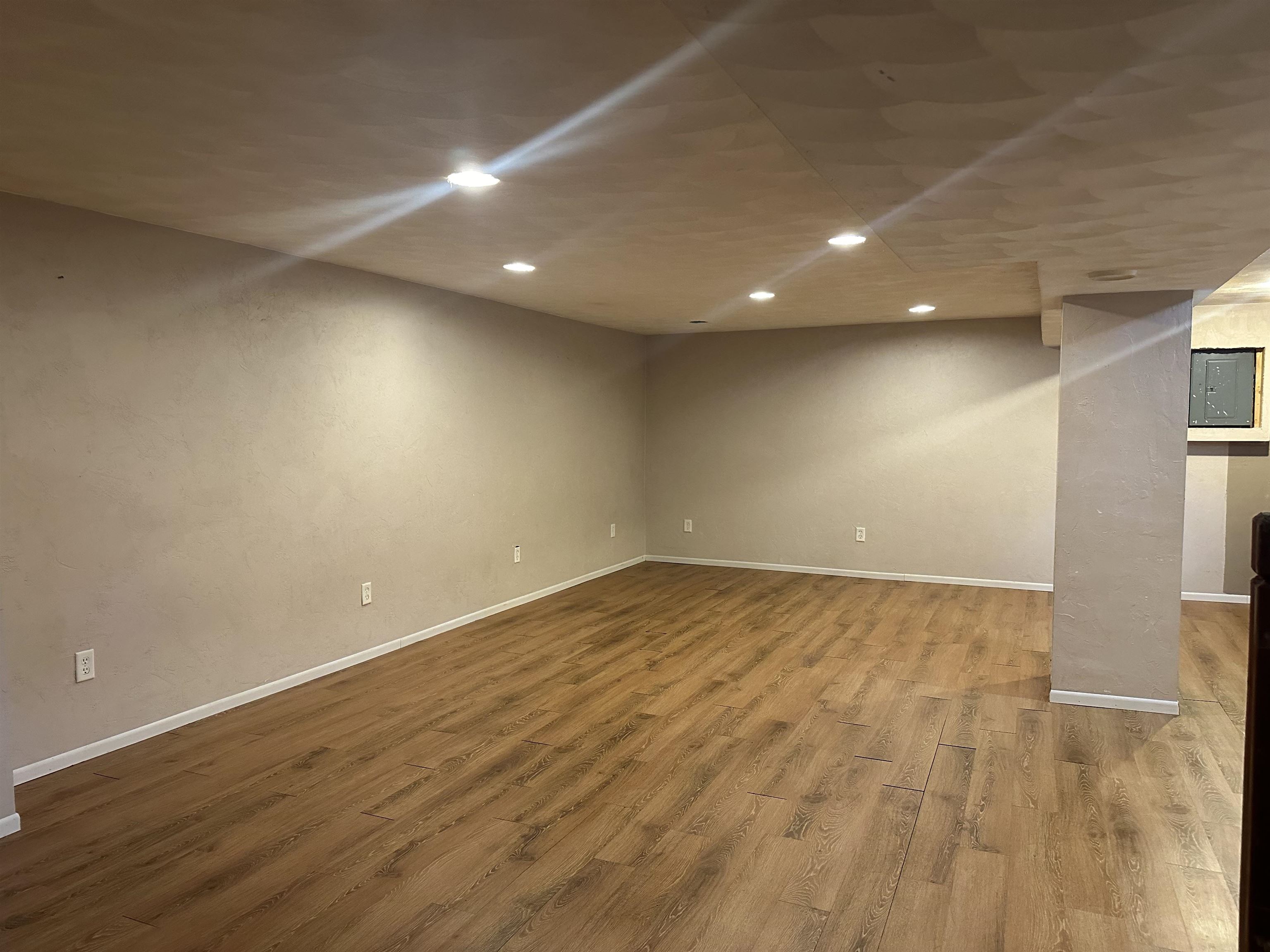 Empty basement room with wood-look flooring, beige walls, recessed lights, and a support column.
