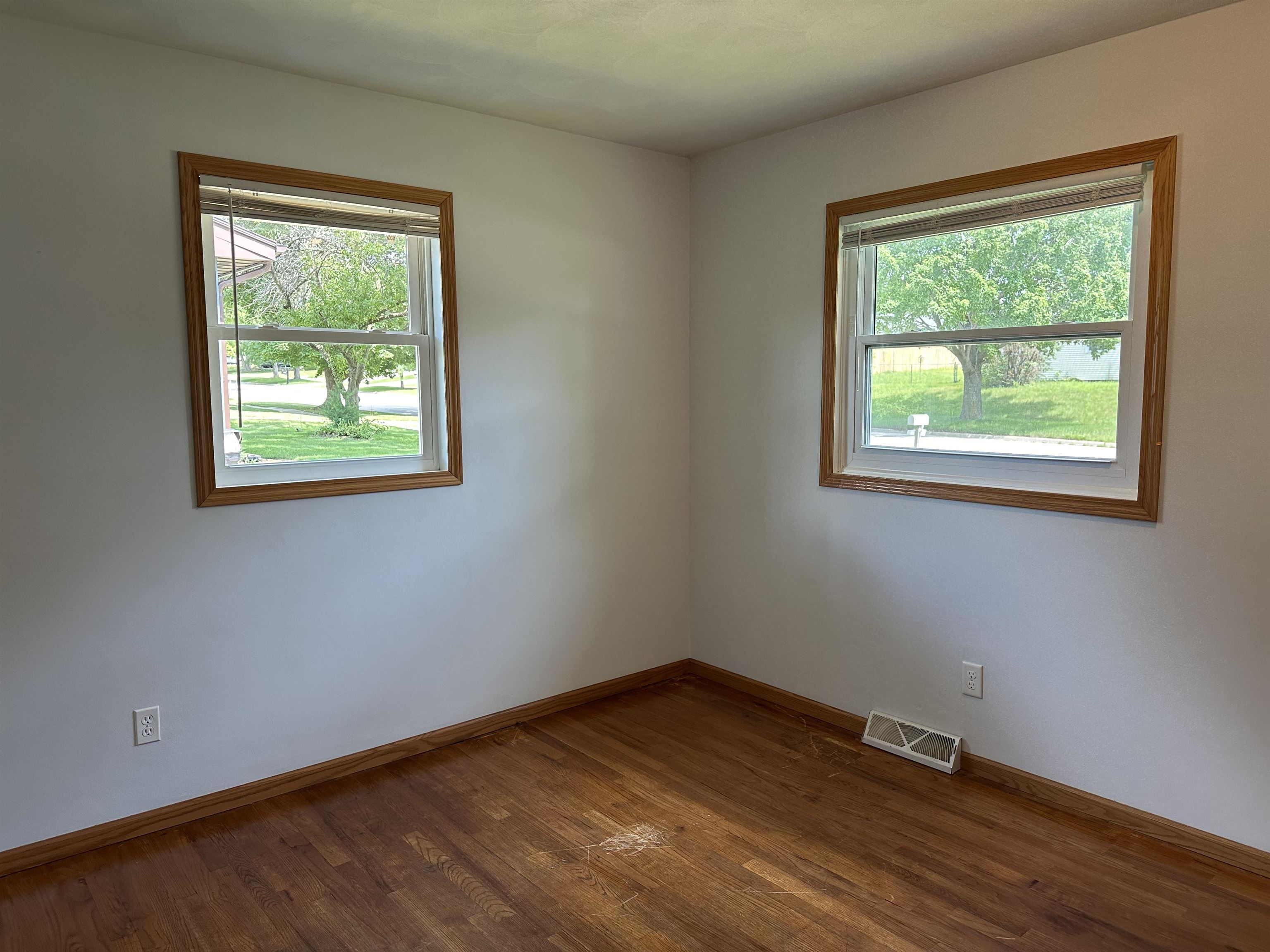 Empty room with worn hardwood floors and two windows overlooking leafy trees.