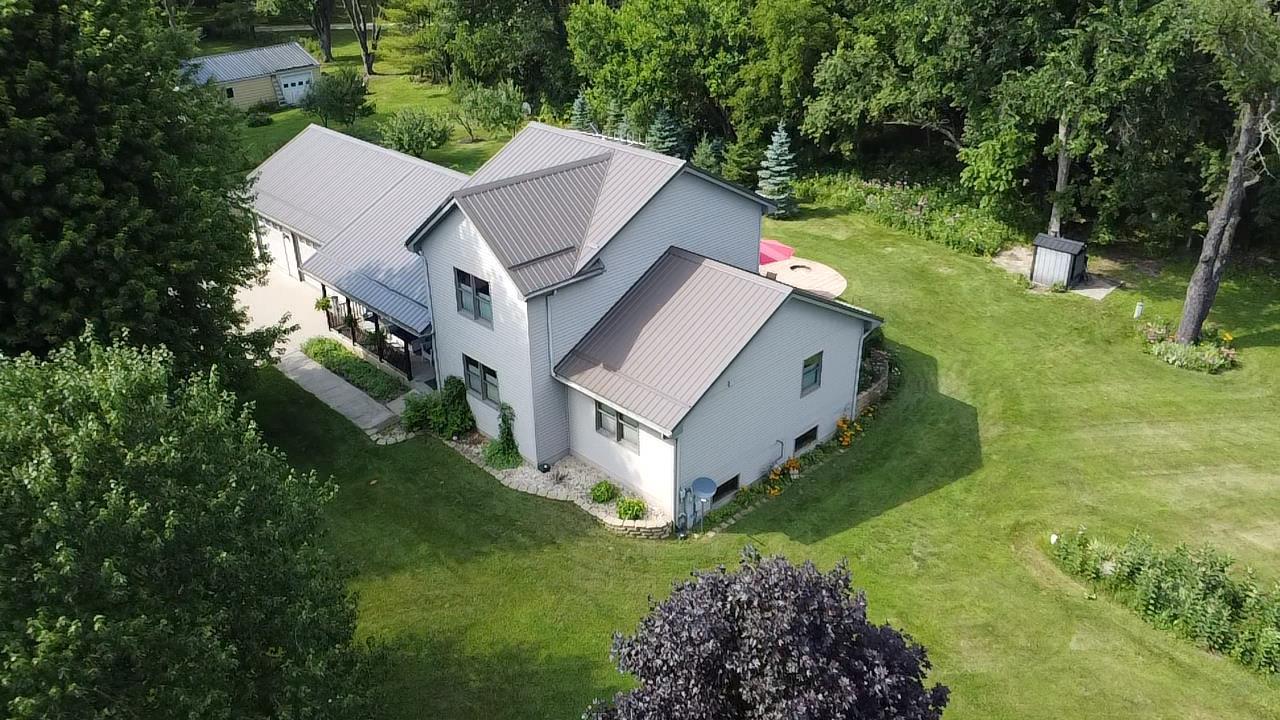 Aerial view of a light-colored home with a multi-section metal roof, broad lawn, and mature trees.