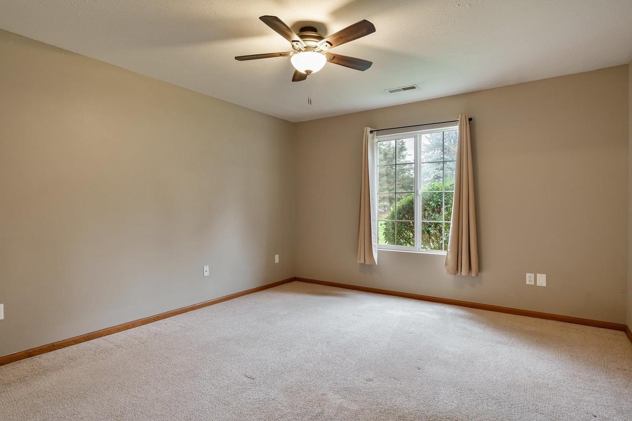 Empty carpeted bedroom with a ceiling fan and a curtained window overlooking greenery.