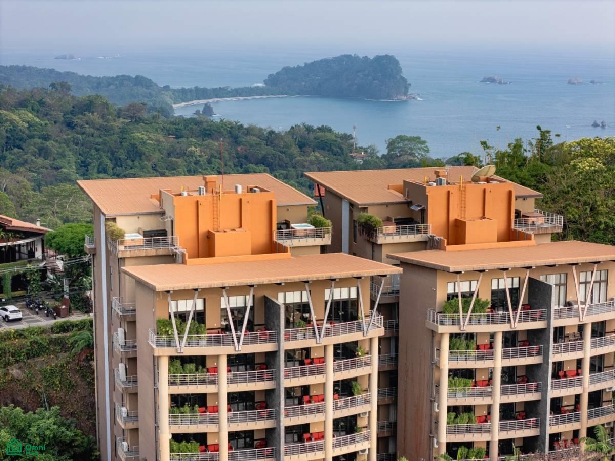 Condominium buildings with orange rooftop structures overlook a tropical bay and forested headland.