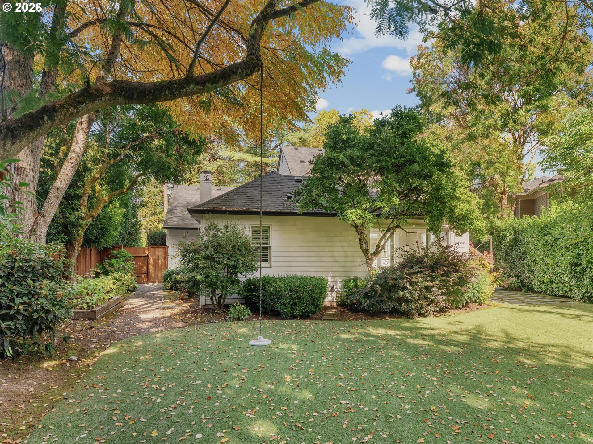 House partly screened by mature trees, with a leaf-strewn lawn and wooden garden gate.