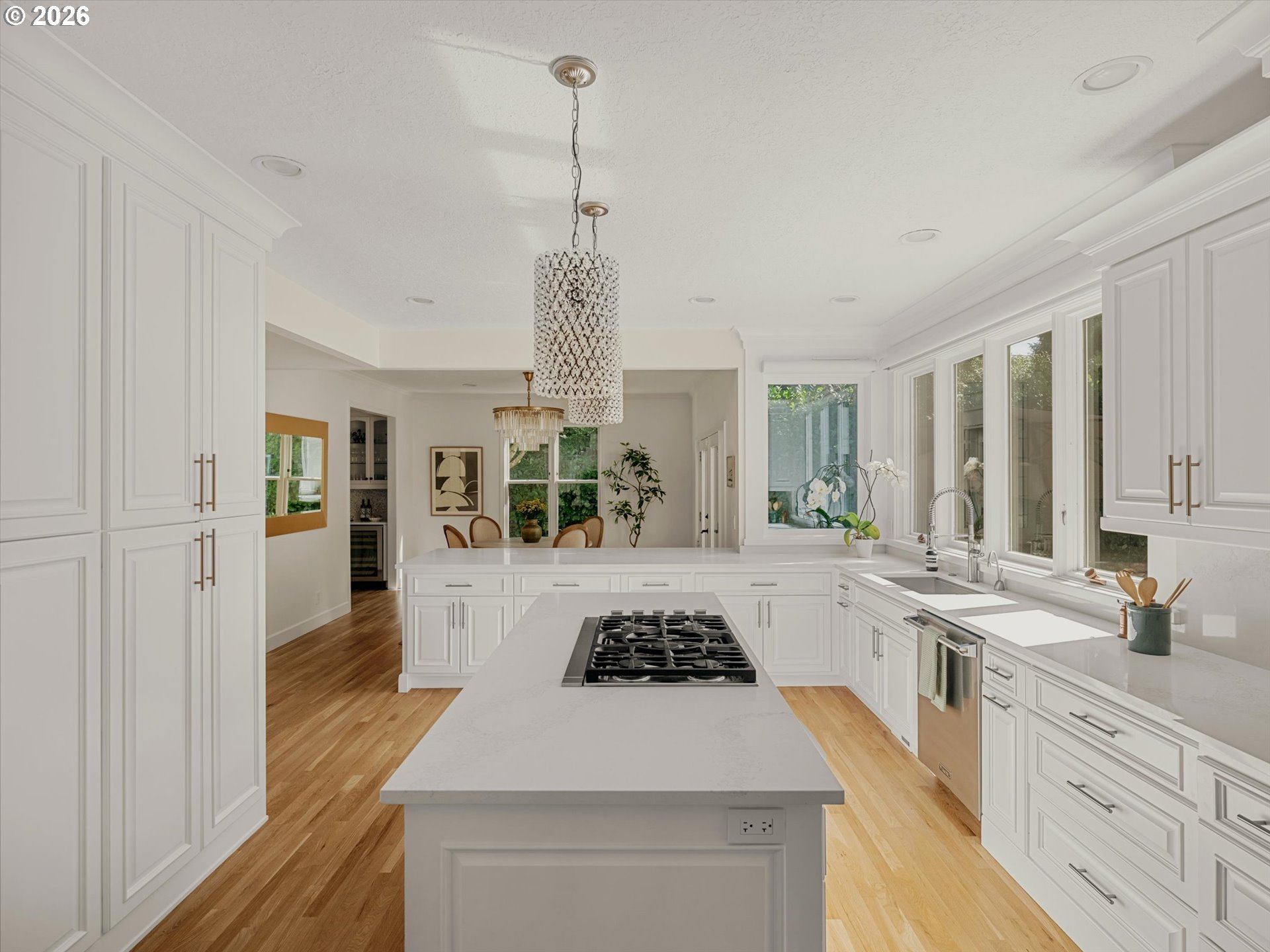 Bright white kitchen with a large cooktop island, hardwood floors, and wide windows above the sink.