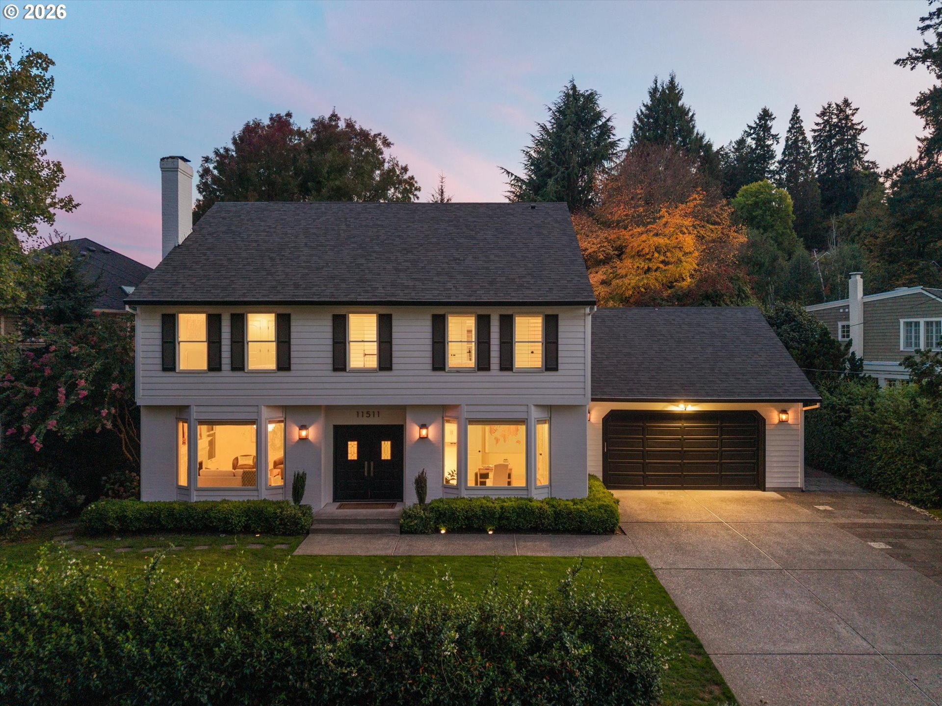 Two-story gray house with warmly lit windows, black double garage door, and autumn trees at dusk.