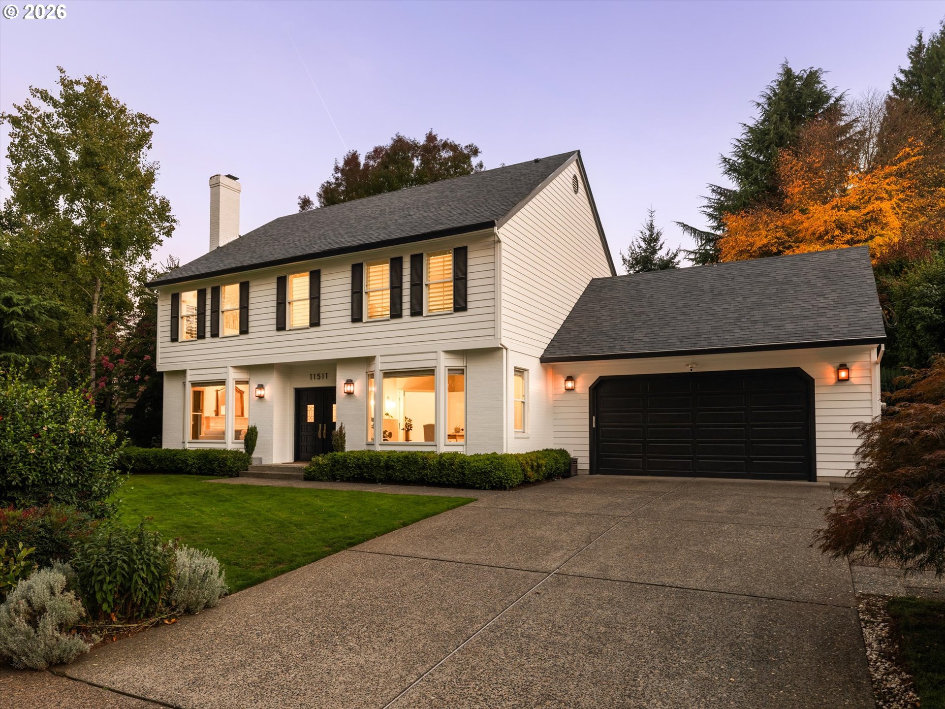Two-story white house with black shutters, a double garage, and autumn foliage at dusk.
