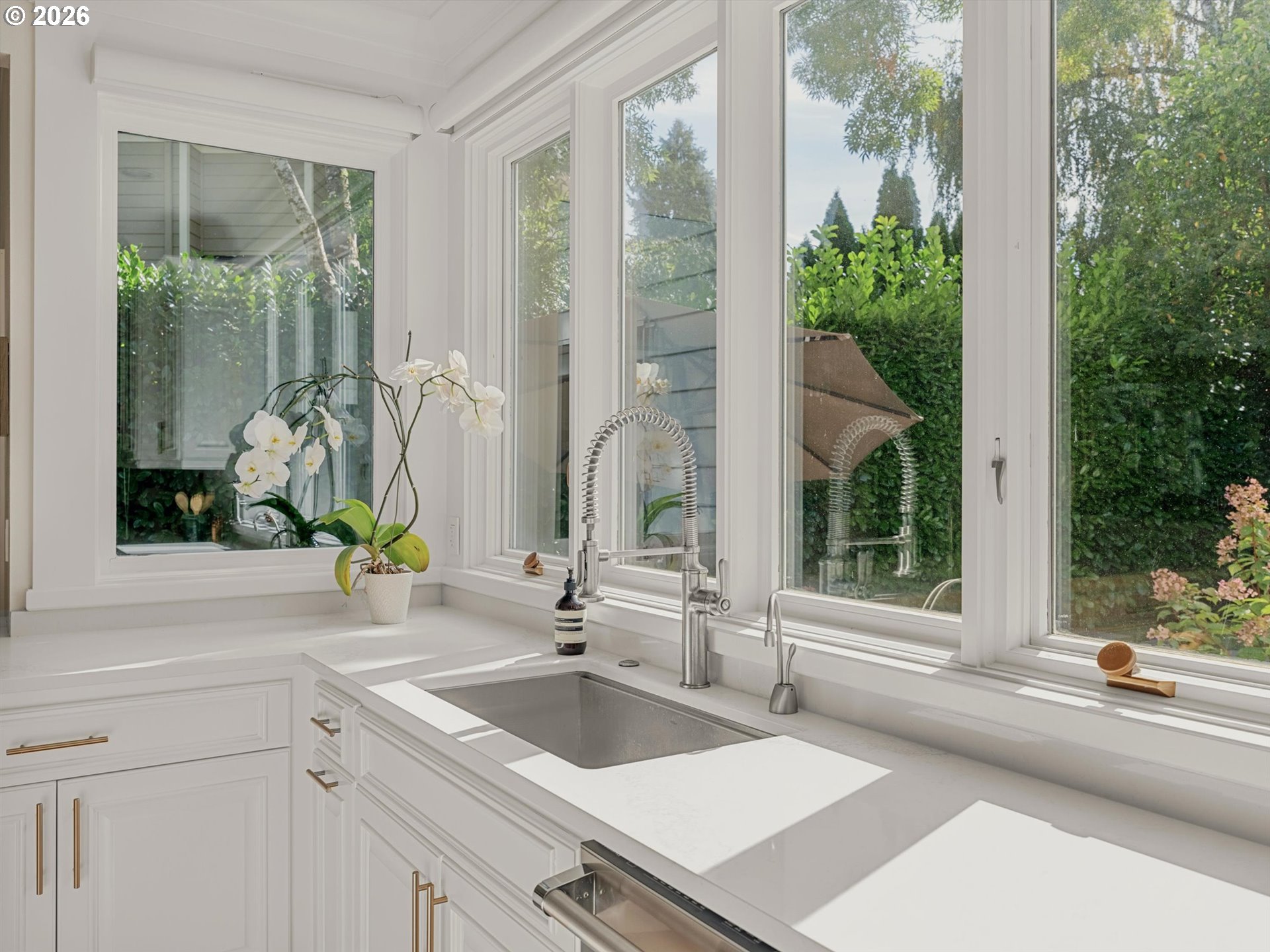 Bright white kitchen sink beneath large windows, with a spring-coil faucet and leafy garden views.