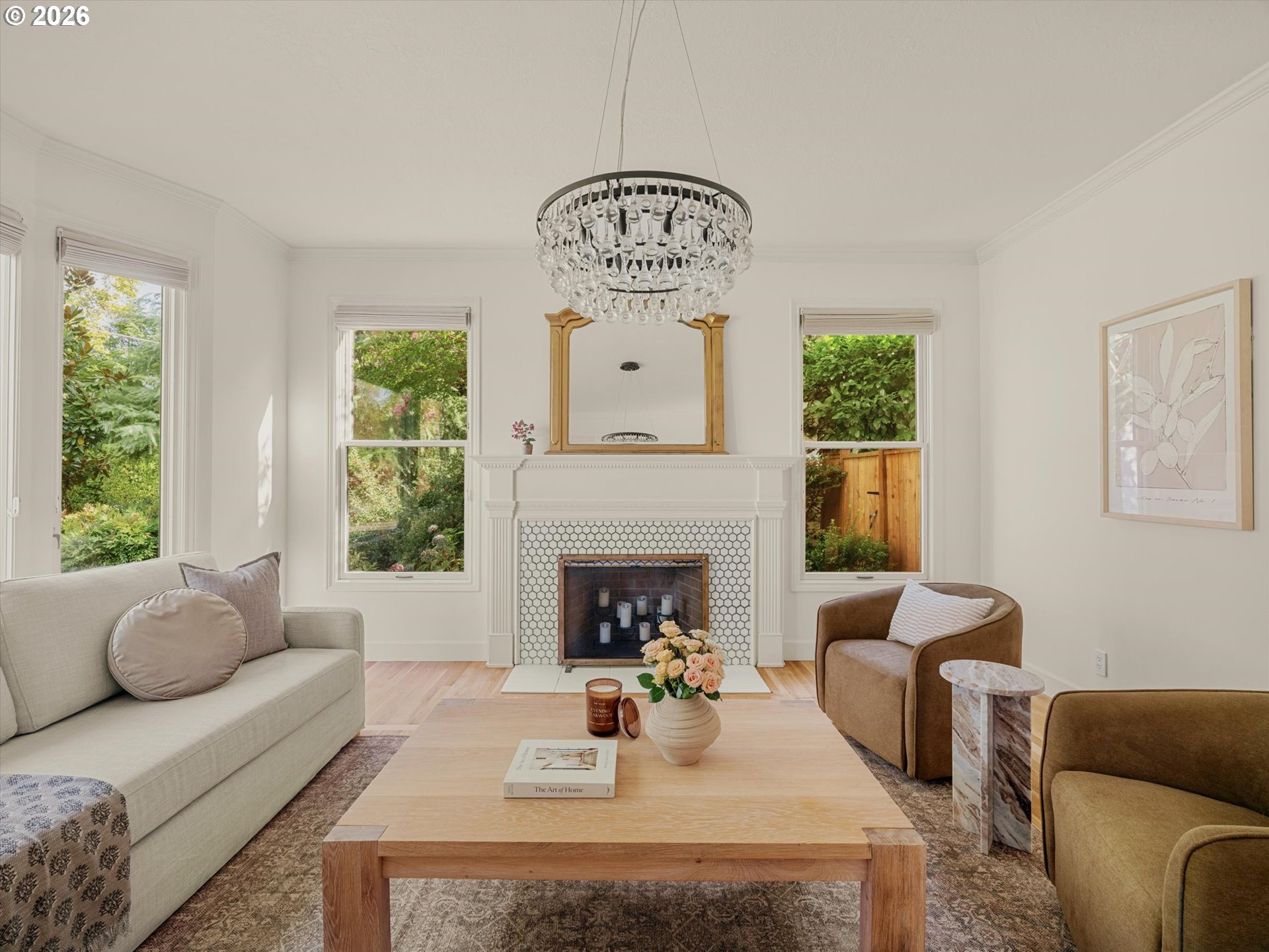 Living room with a white tiled fireplace, crystal chandelier, and windows overlooking greenery.