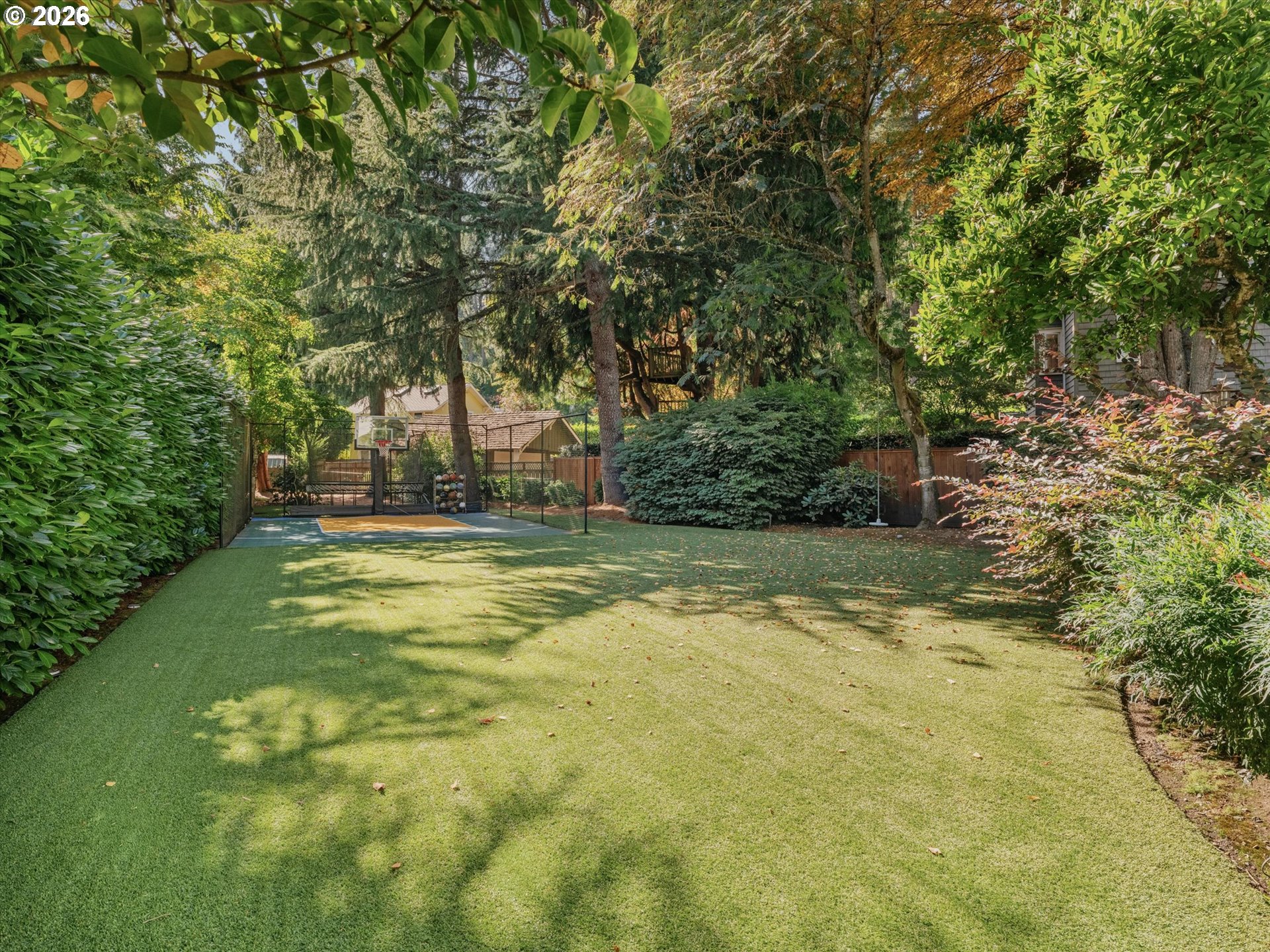 Landscaped backyard with a broad lawn and a fenced basketball court beneath mature trees.