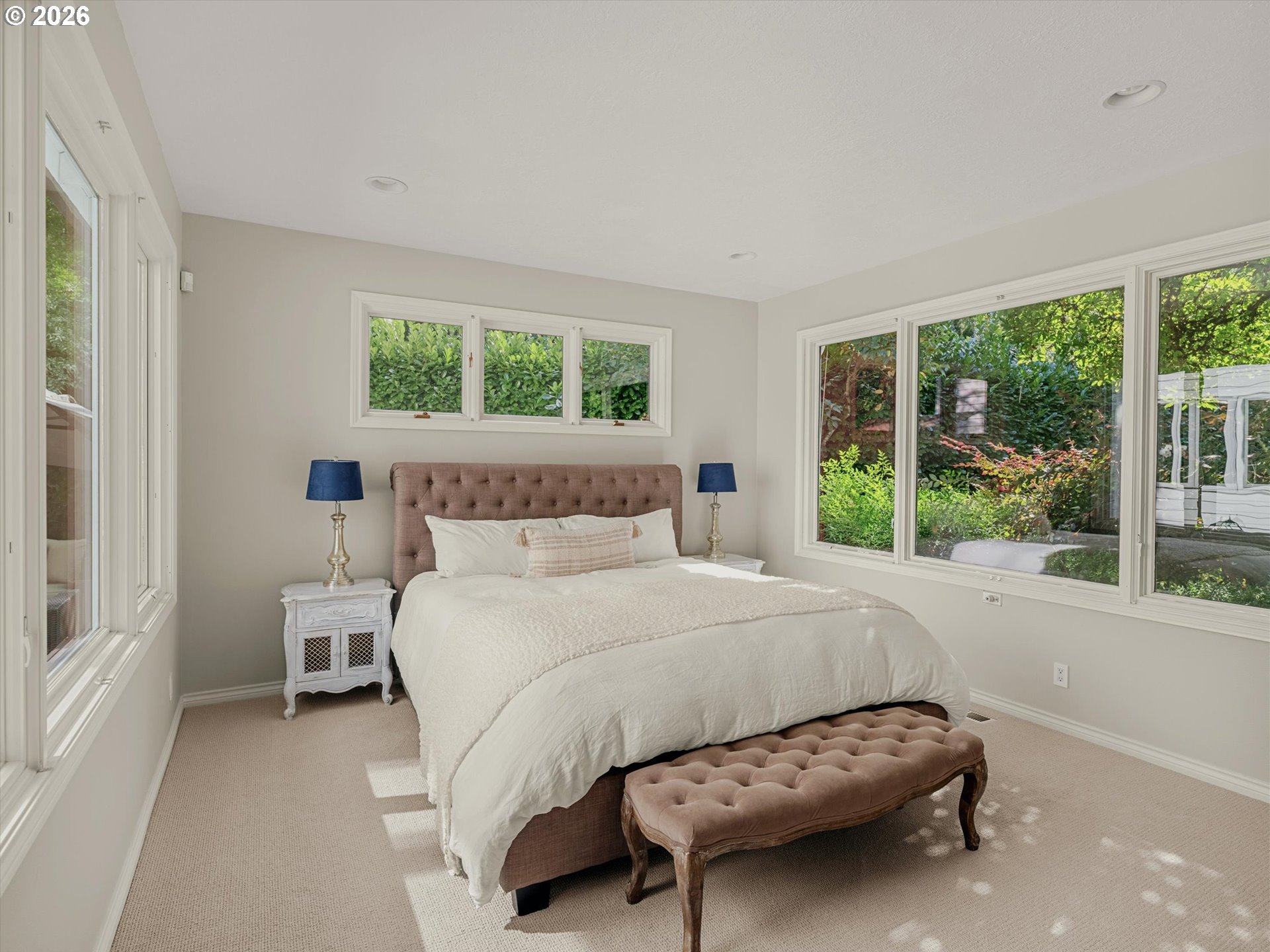 Furnished bedroom with white bedding, a tufted headboard, and large windows overlooking greenery.