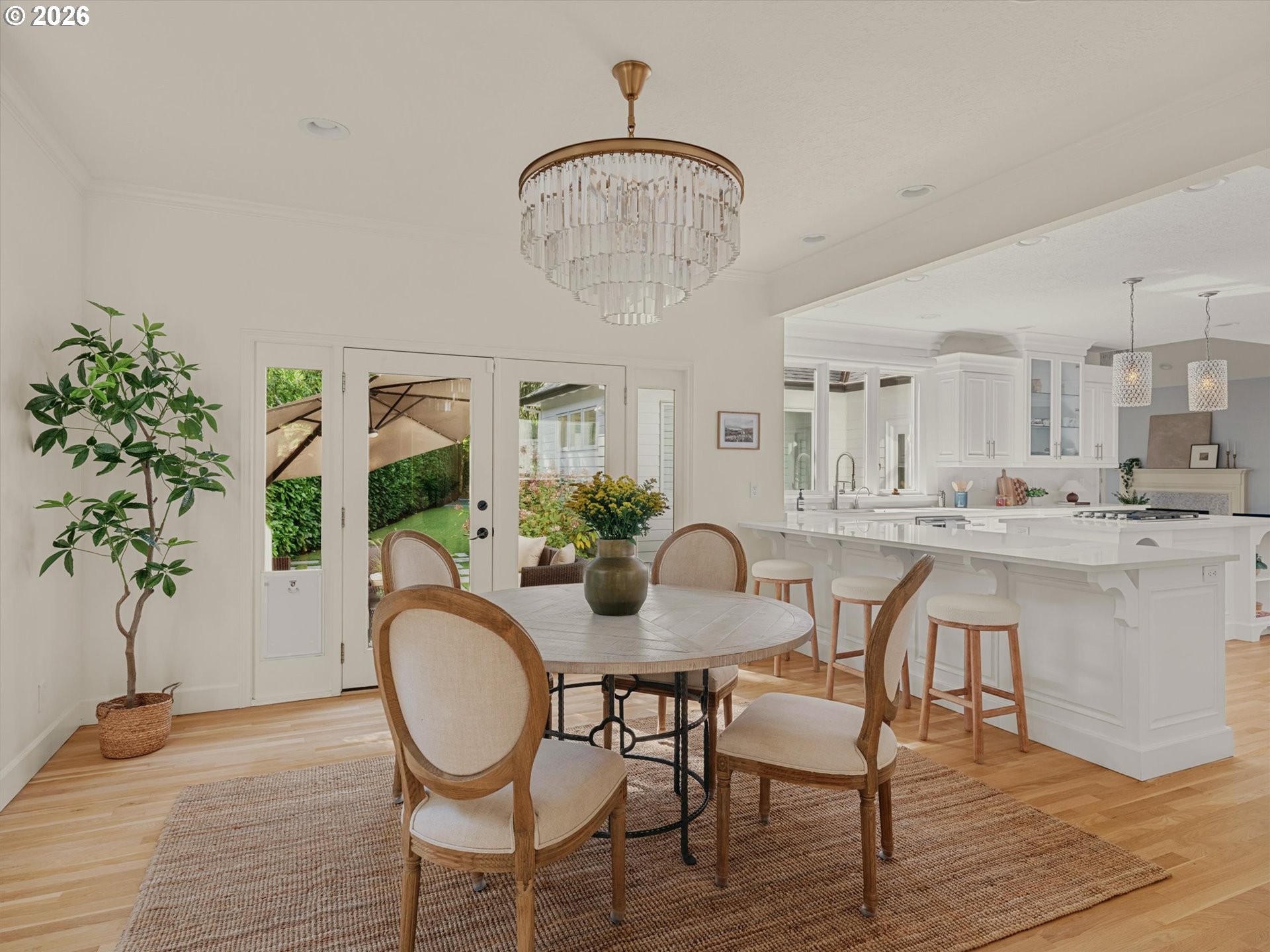Dining area with a round table beneath a crystal chandelier, adjoining a bright white kitchen.
