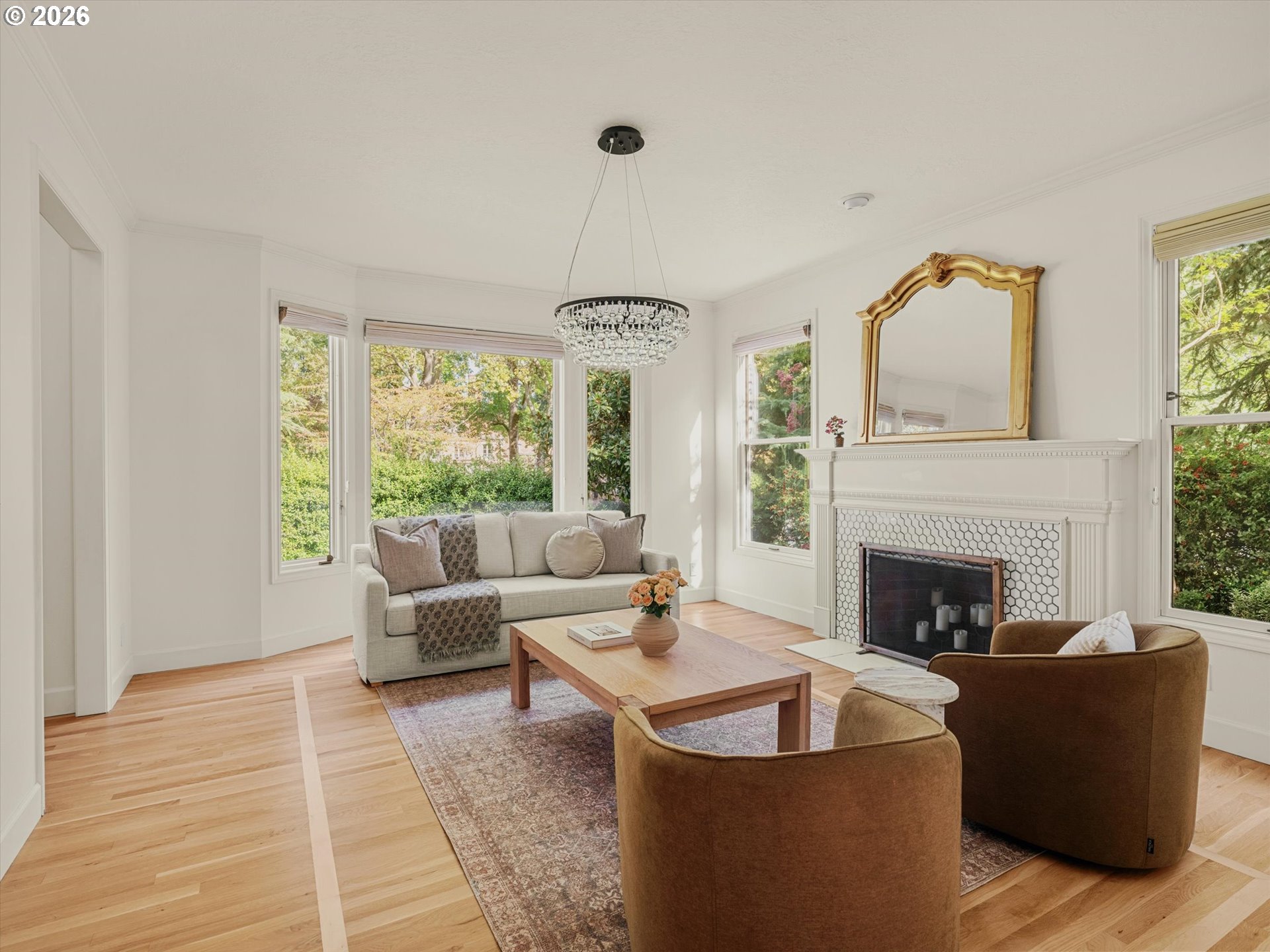 Bright living room with a white tiled fireplace, crystal chandelier, and large garden-facing windows.