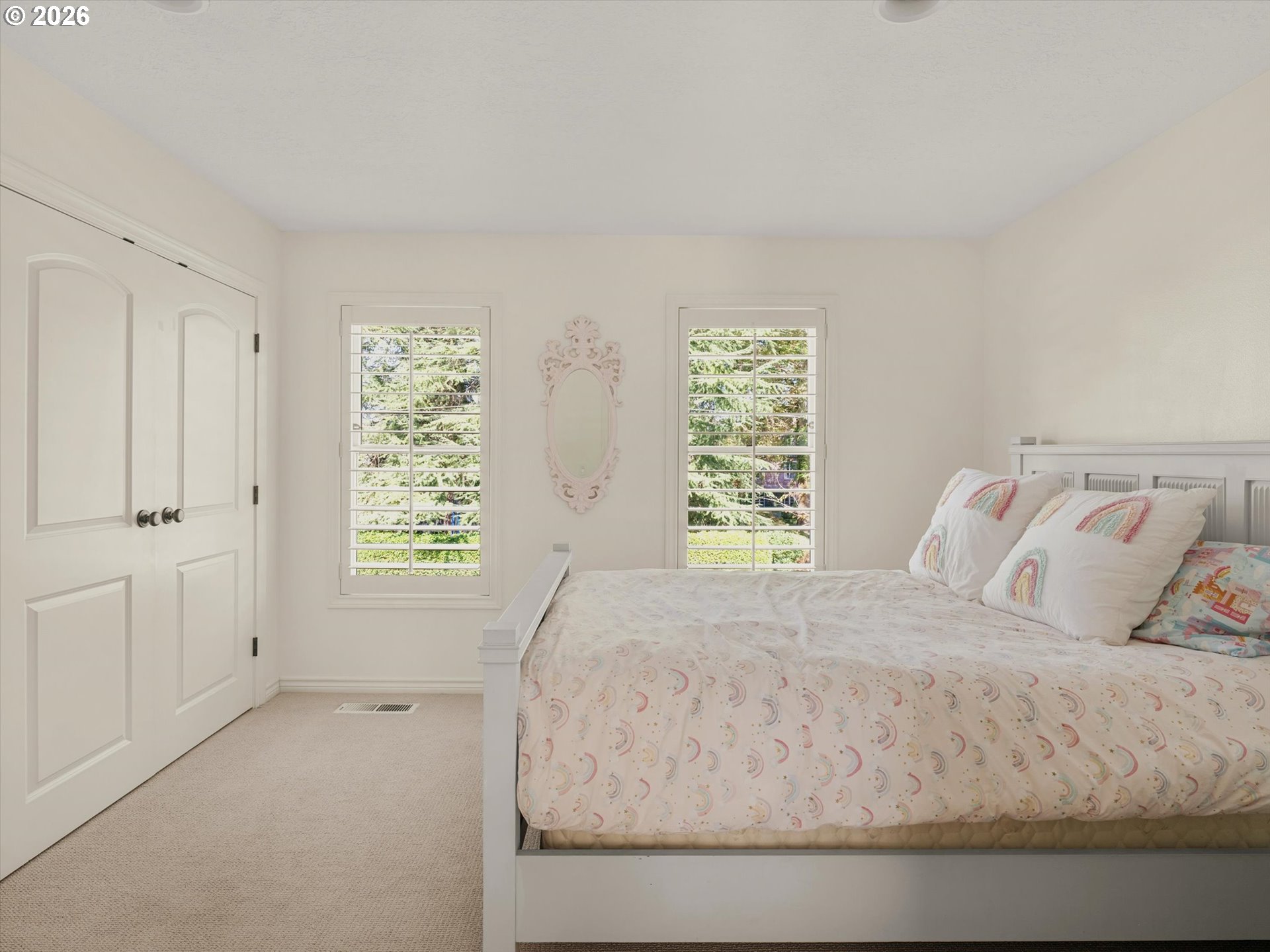 Bright bedroom with a white bed, pastel rainbow bedding, and two shuttered windows.